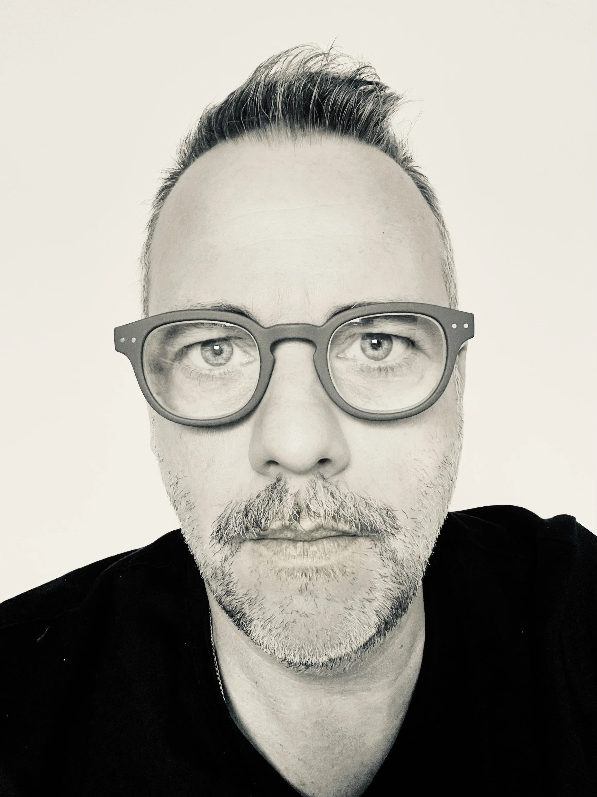 A black and white photo of a middle-aged man with light-colored facial hair, wearing large glasses, with short styled hair on top, and a serious expression, dressed in a dark jacket.