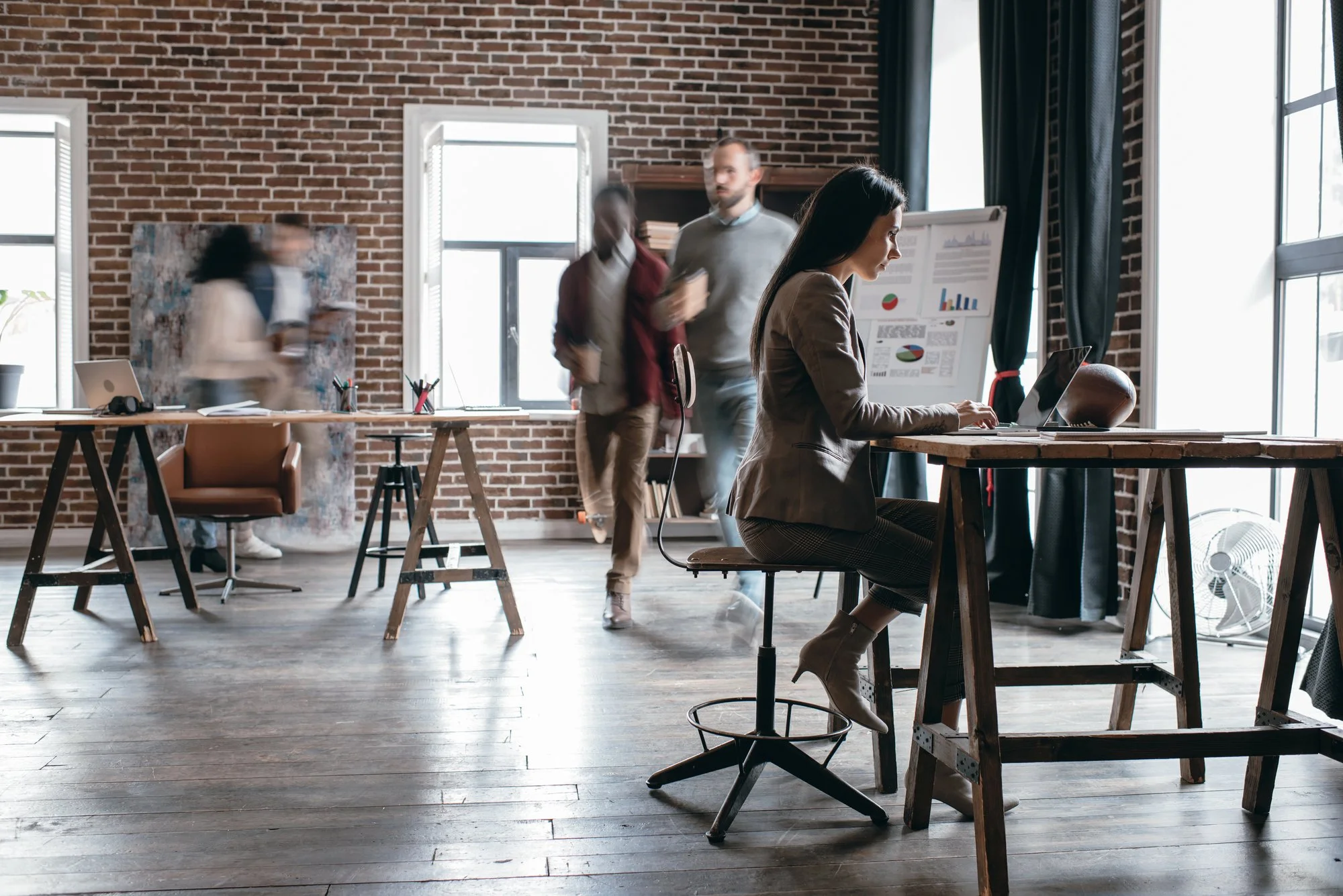 A woman working on a laptop at a rustic wooden desk in an industrial-style office with exposed brick walls, large windows, and modern furniture. Four people are walking in the background, blurry.