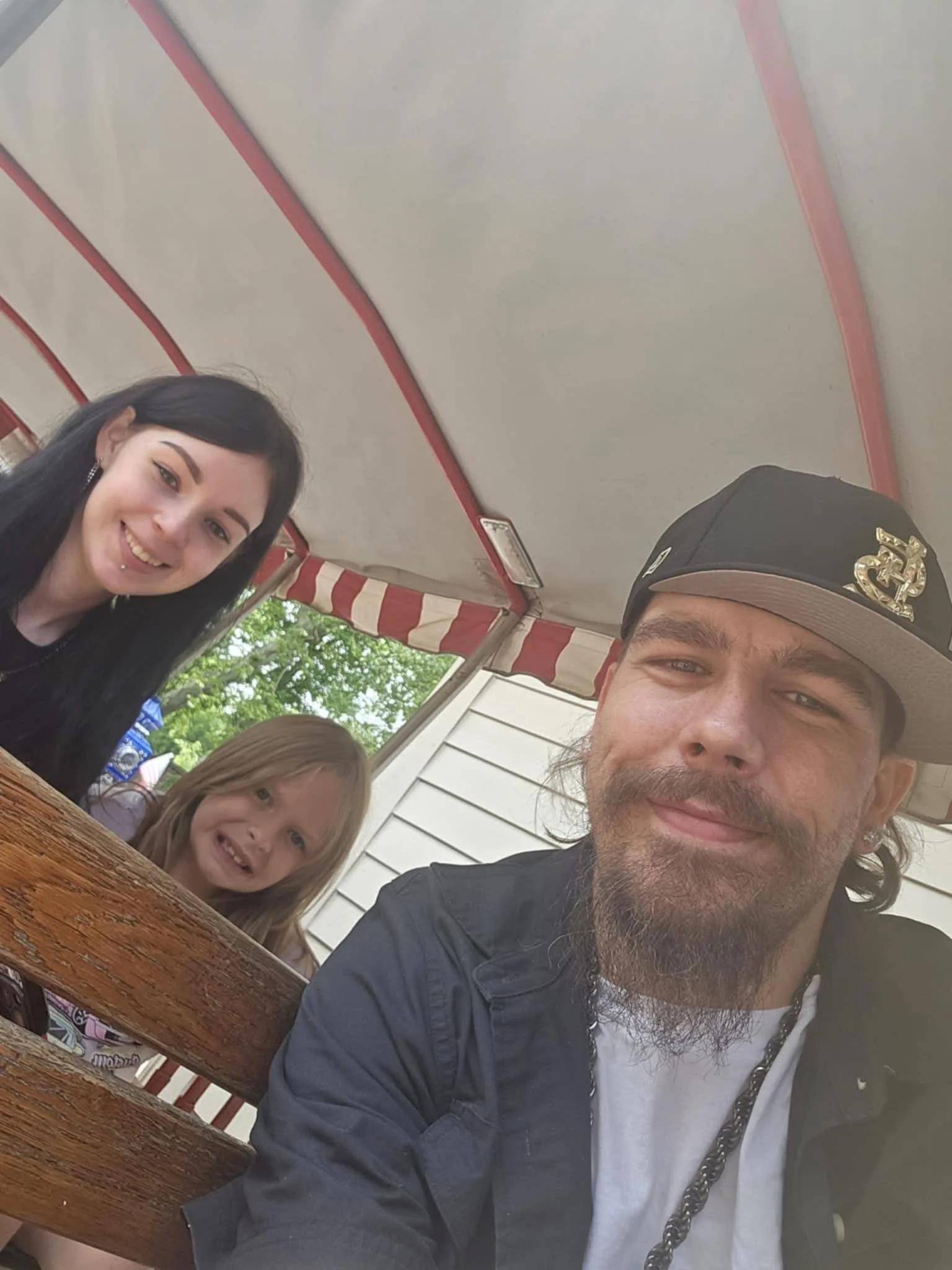 A man wearing a black cap and a woman with black hair and earrings sitting at a wooden outdoor table with a young girl with blonde hair. They are outdoors under a red and white striped canopy, smiling at the camera.