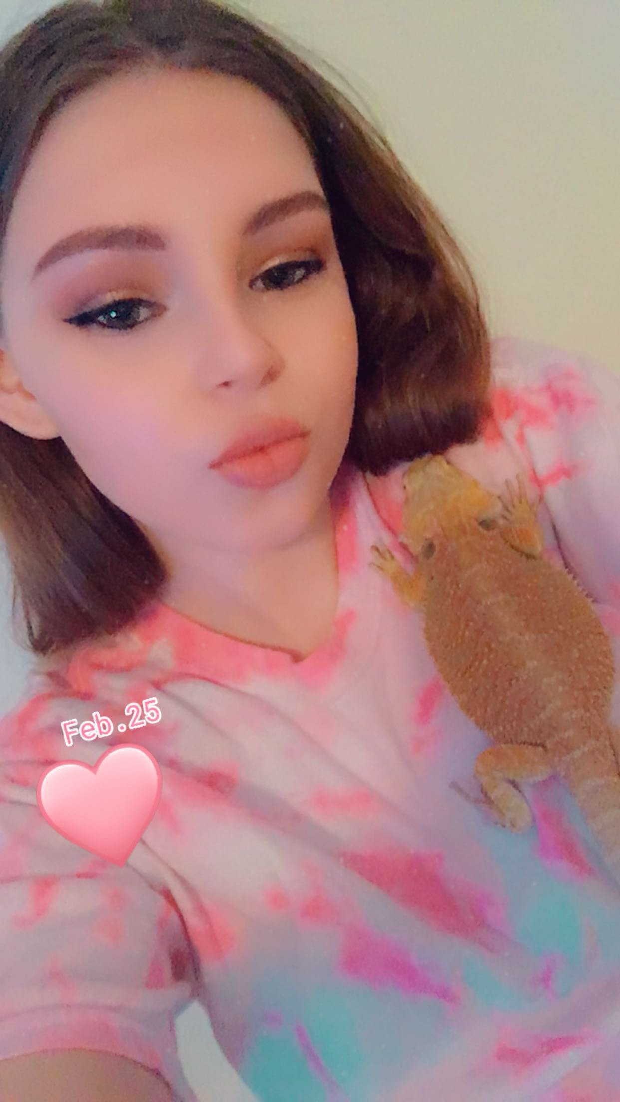 A young woman with makeup and shoulder-length brown hair taking a selfie with an orange bearded dragon sitting on her chest. She is wearing a pastel tie-dye shirt with pink, purple, and blue colors. There is a pink heart sticker with the text "Feb. 25" on it.