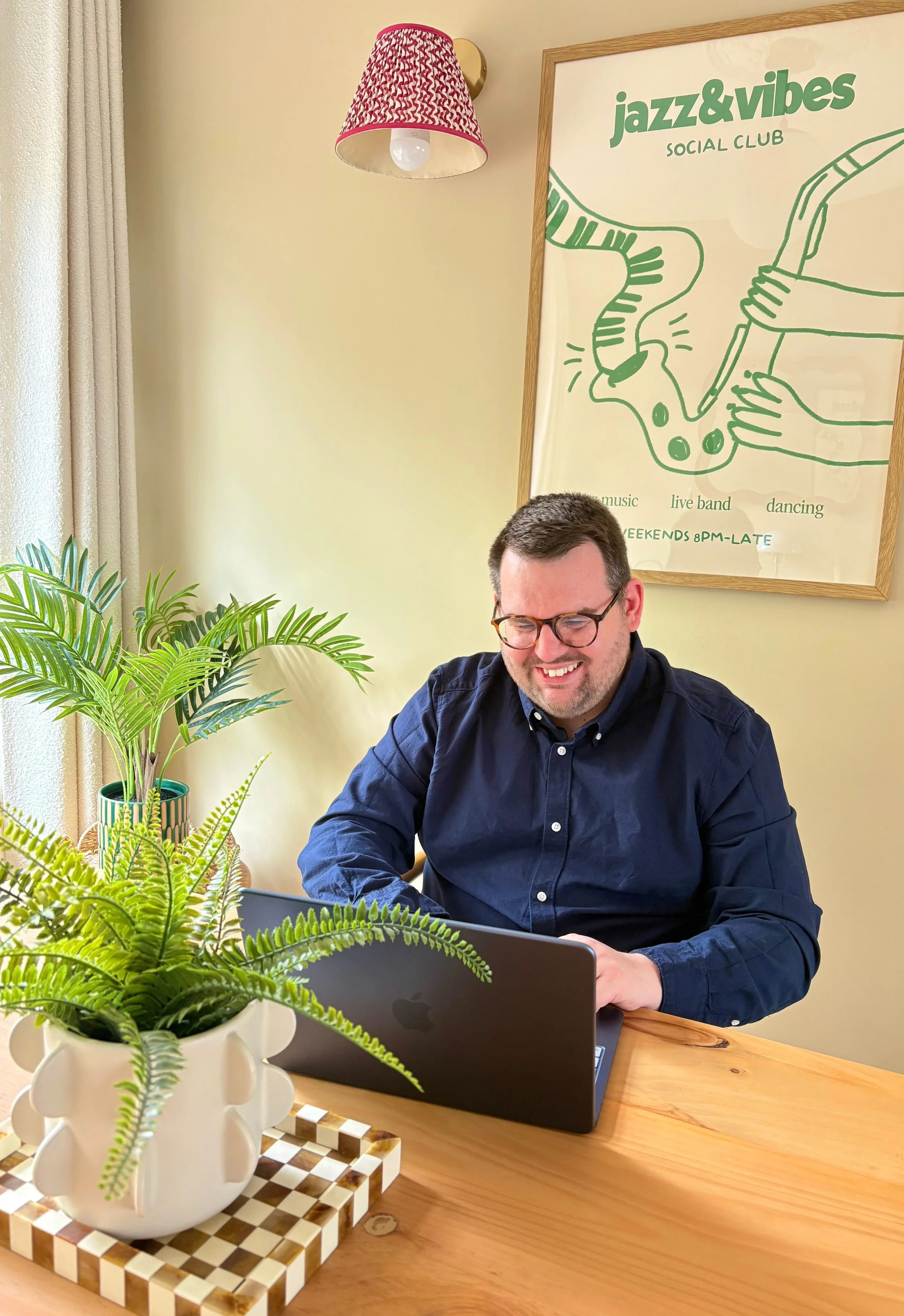 A man with glasses wearing a dark blue shirt sitting at a wooden table with a laptop in front, smiling while looking at the screen. There are green houseplants on the table and a framed poster on the wall behind him that says 'jazz&vibes SOCIAL CLUB' with illustrations of hands holding a saxophone and a microphone. A pink patterned lampshade hangs above, and beige curtains are visible on the side.