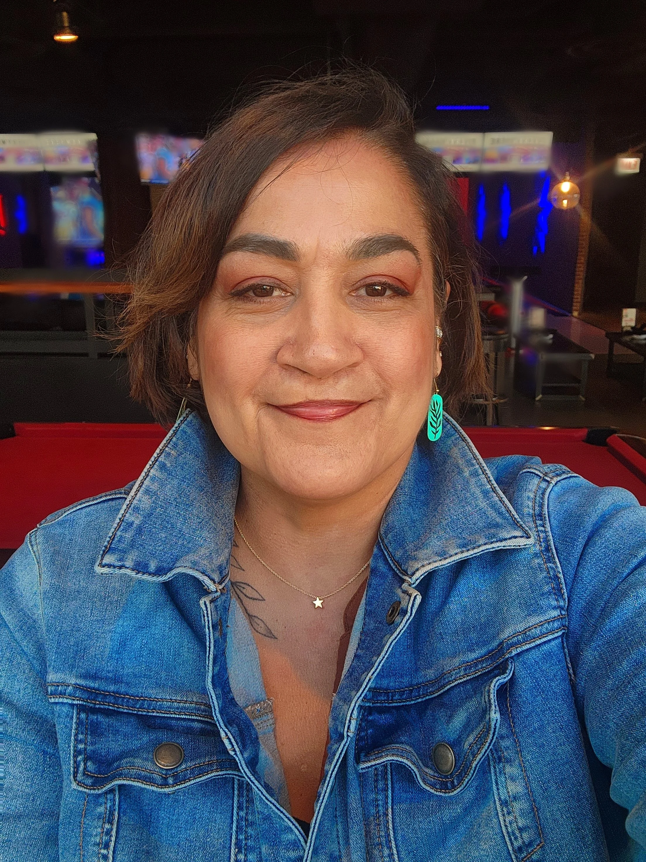 A woman with short dark hair wearing a denim jacket and turquoise earrings, smiling at the camera in a dimly lit bar or entertainment venue with screens and colorful lights in the background.