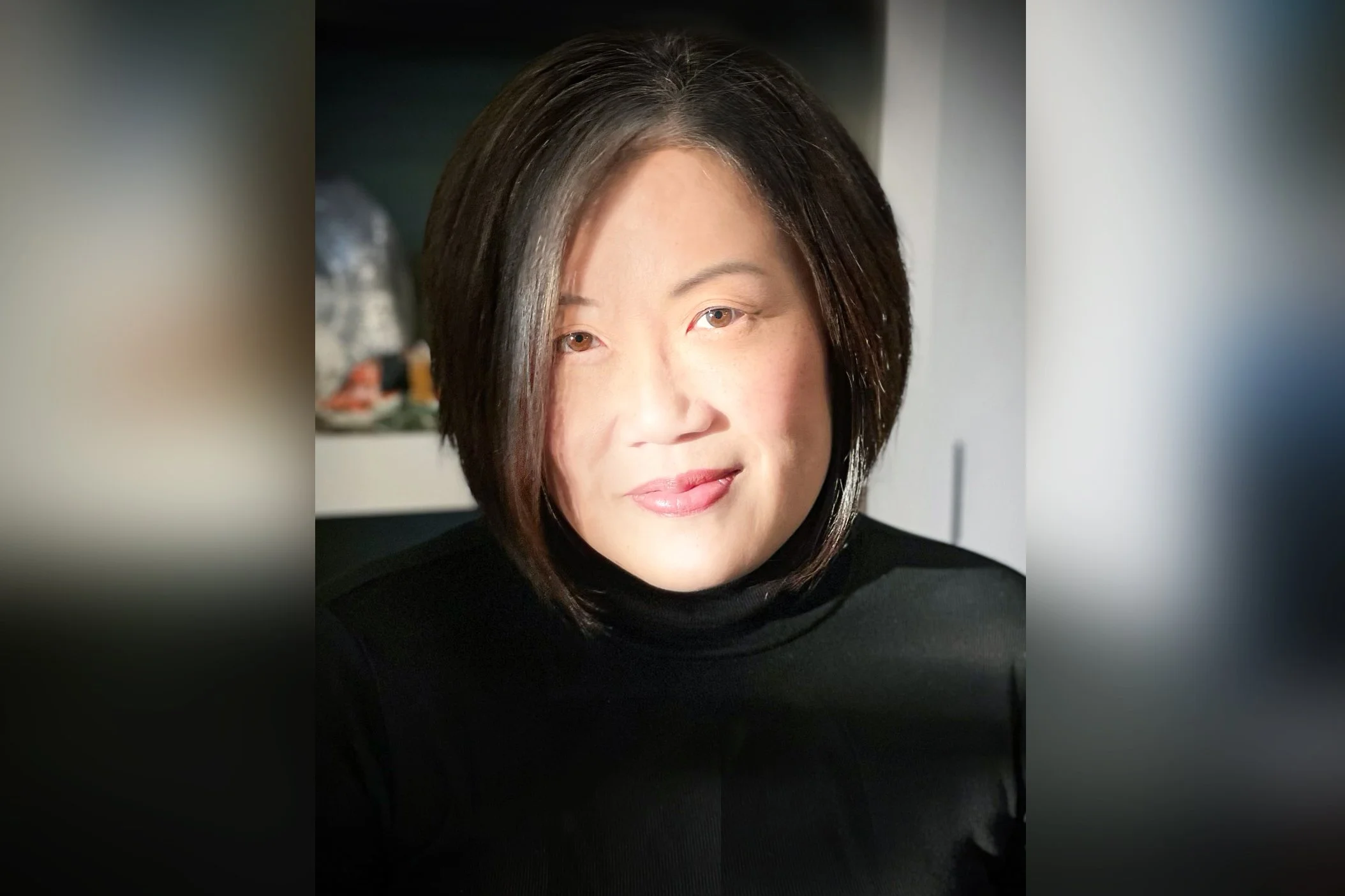 A woman with shoulder-length dark brown hair, wearing a black turtleneck, looking at the camera with a slight smile in a well-lit indoor setting.