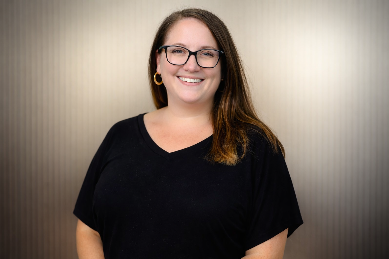 A woman with long brown hair, wearing black glasses, gold hoop earrings, and a black V-neck shirt, smiling at the camera against a gray, striped background.