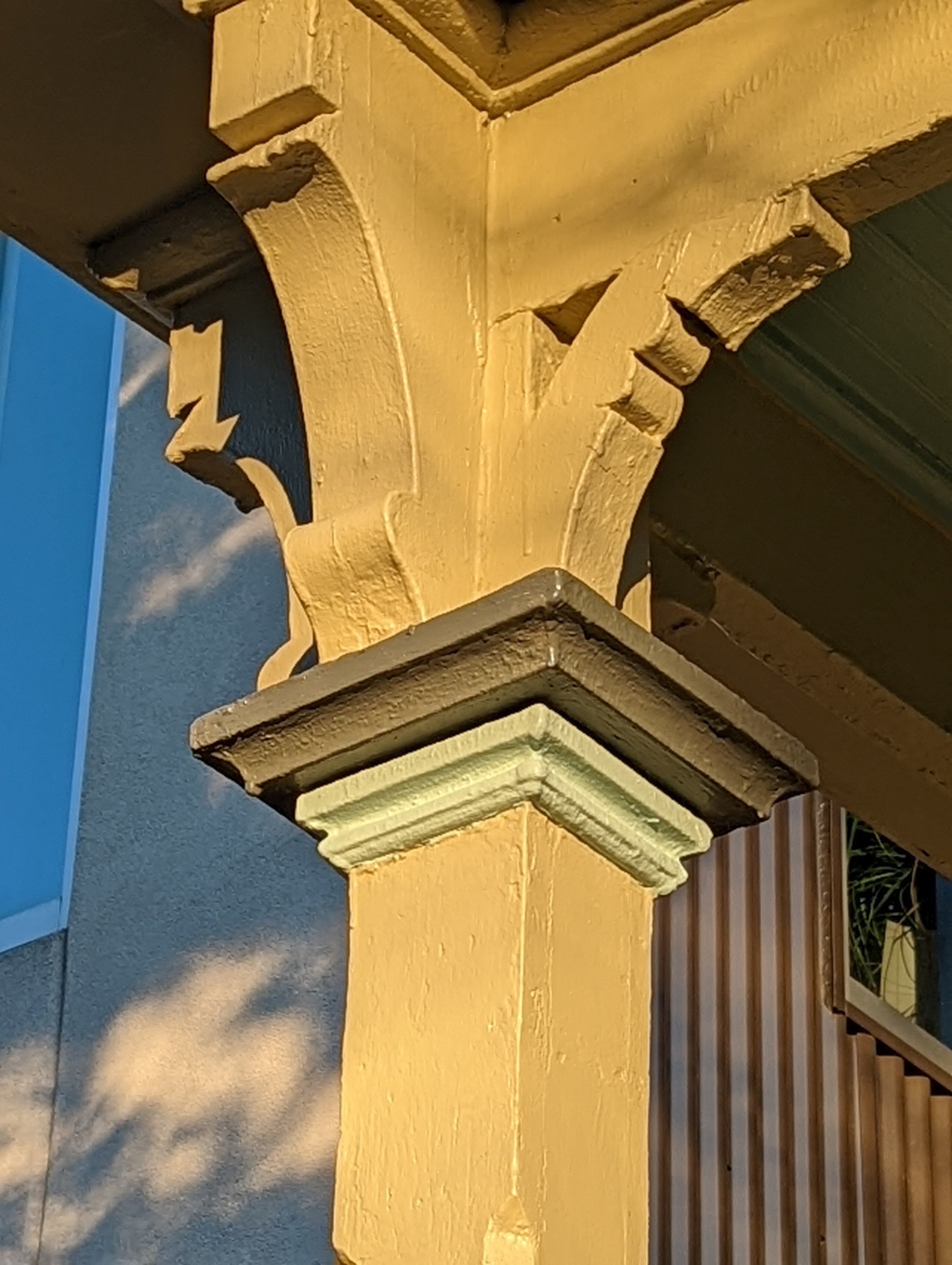 Close-up of decorative architectural corbels painted yellow, part of a building or structure, with a shadow cast on a gray ground and a wooden area in the background.