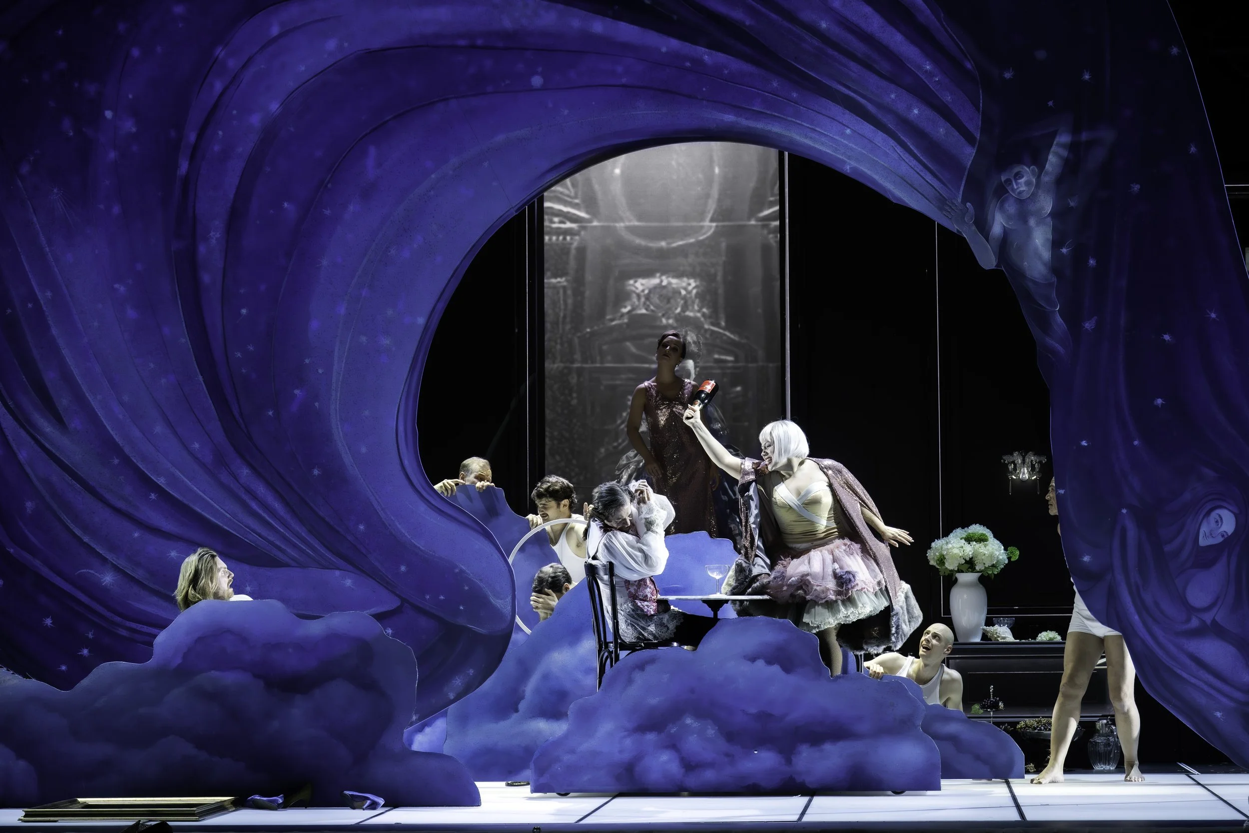 A theatrical ballet scene with dancers on stage, framed by a large blue cloud-like set piece with stars. The performers are dressed in pastel costumes, with some holding props like flowers and bottles, engaging in expressive poses.