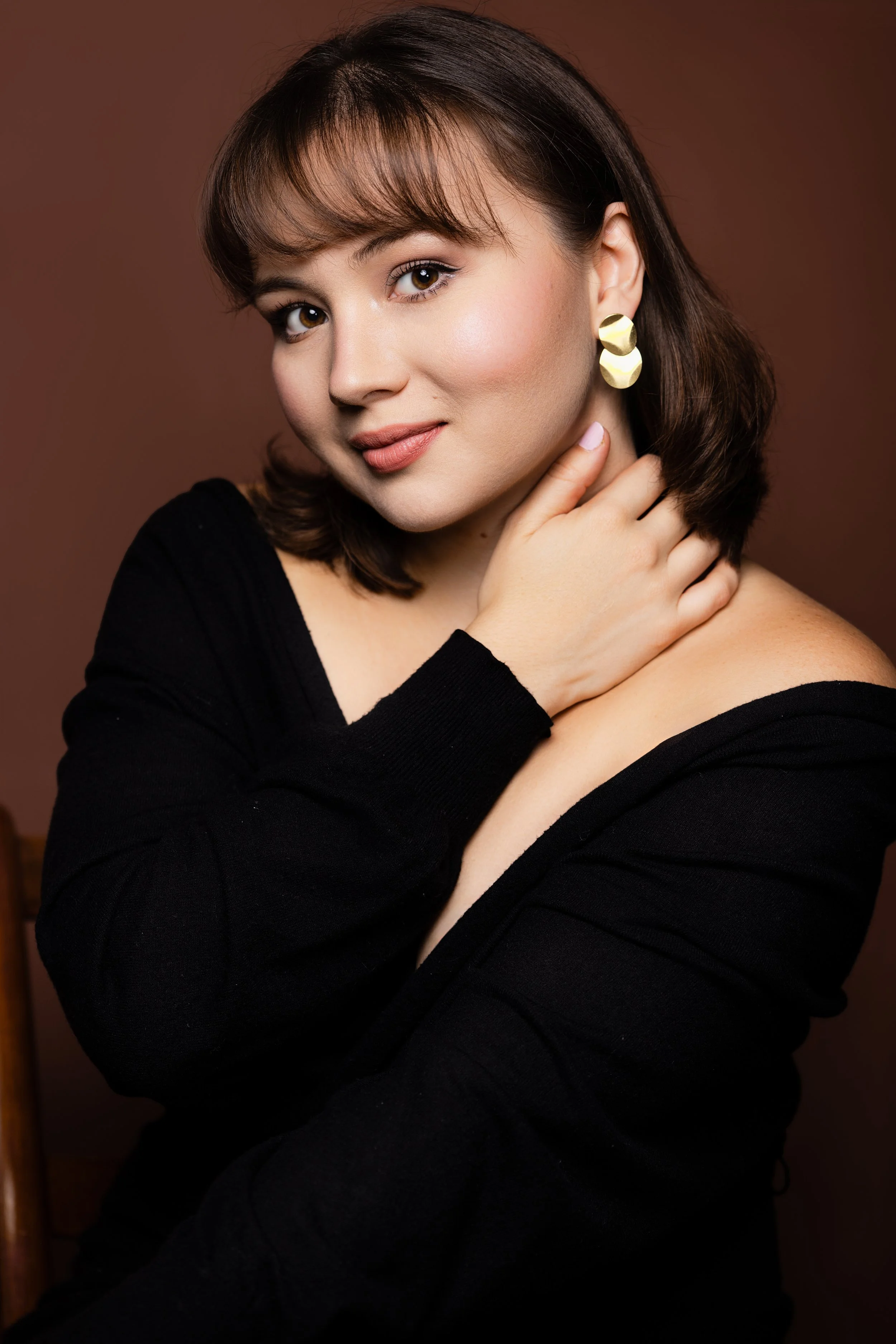 A woman with brown hair, wearing large gold earrings and a black top, touching her neck and shoulder, posing against a brown background.