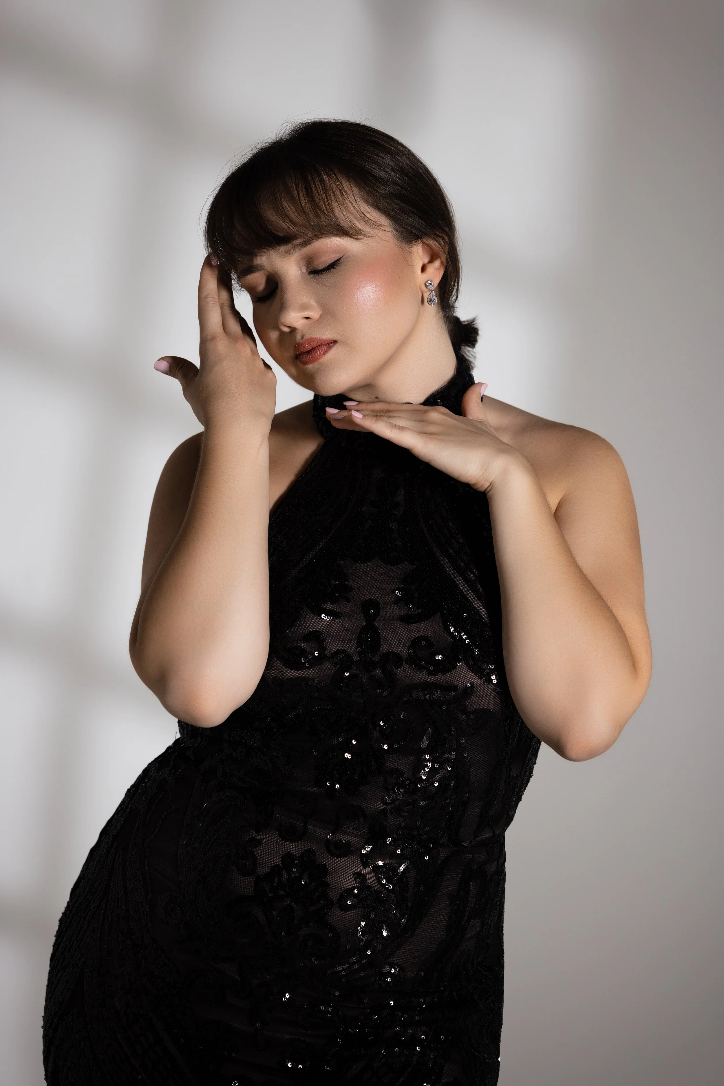 A woman with brown hair styled with bangs, wearing a black dress with sequins, touching her face with her eyes closed and a serene expression.