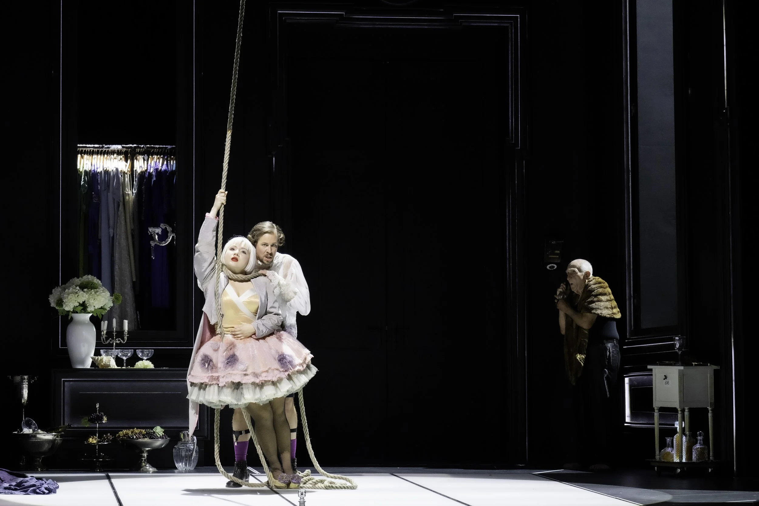 A theatrical stage scene with three actors in elaborate costumes. A woman in a pink tutu and fishnet stockings stands in front, while a man in a light-colored coat is holding her around her neck and holding a rope. In the background, an older woman i