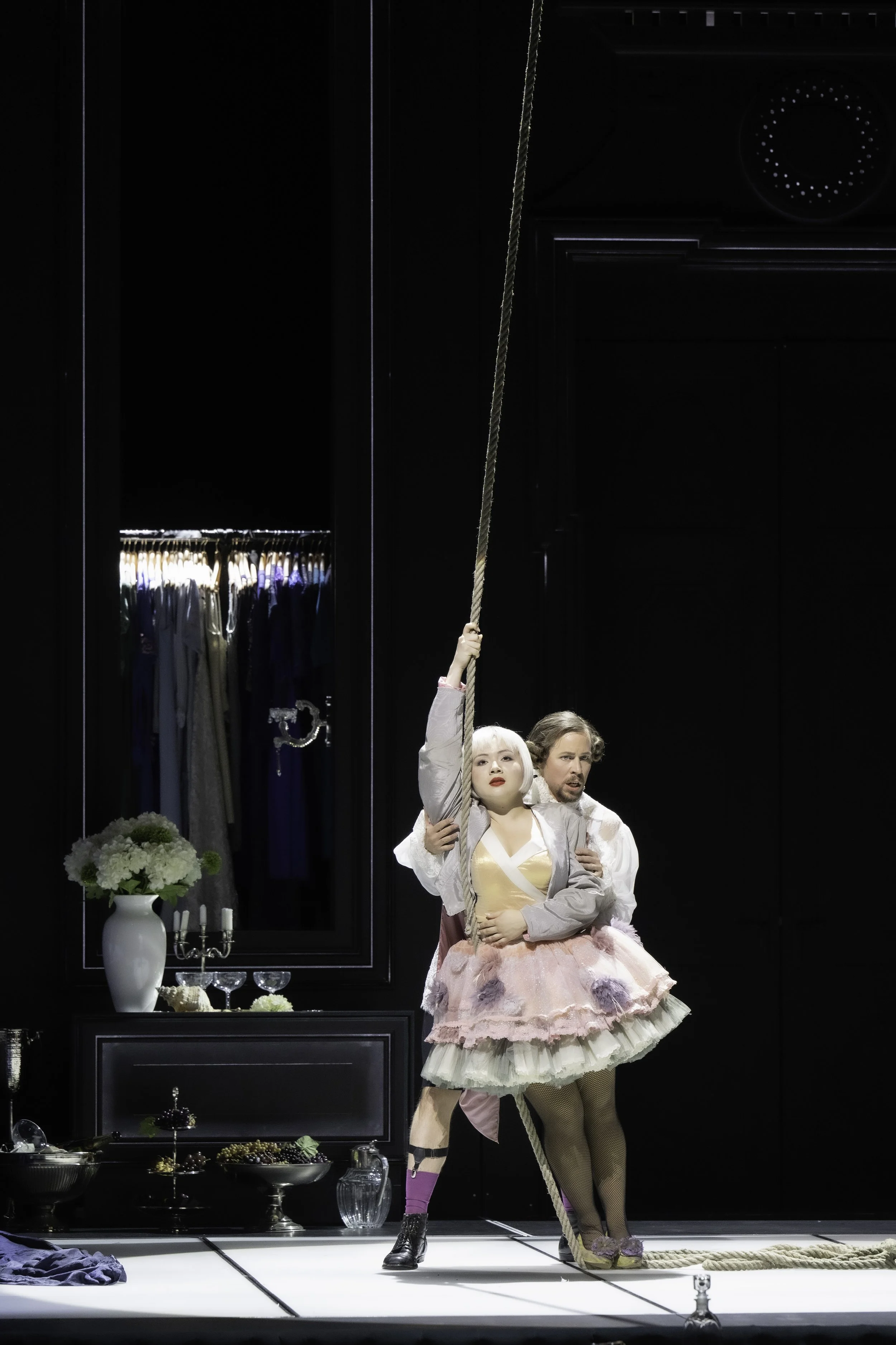 The image shows a theatrical performance scene with three performers on stage, one holding a rope and two standing close together. The stage is set with a black backdrop, a large mirror, and decorative elements such as a white vase with flowers, cand