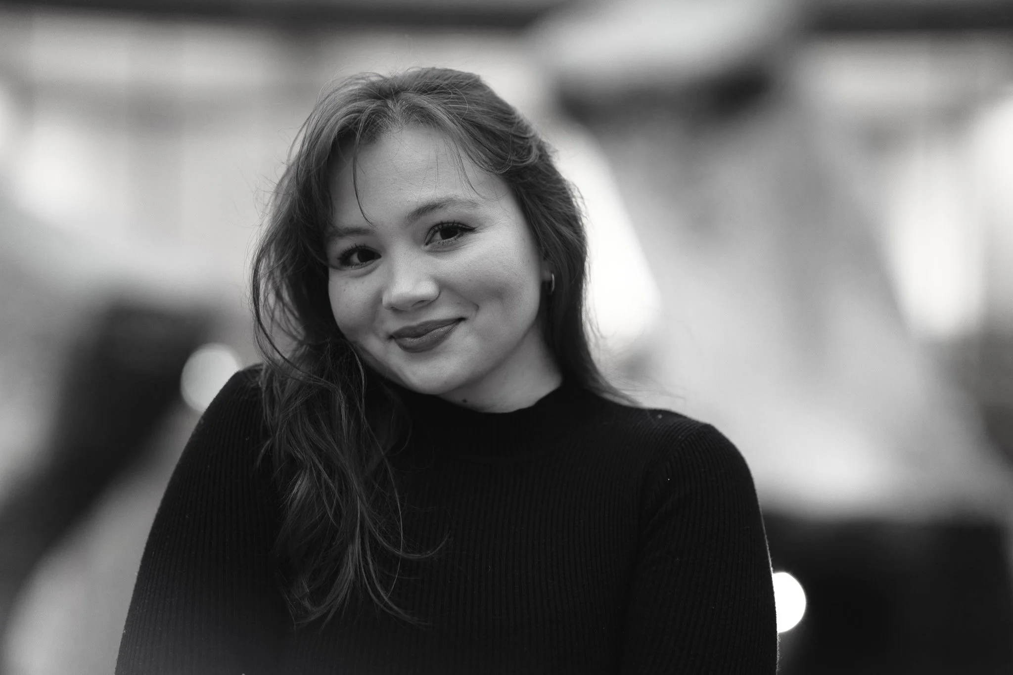 A black and white photo of a young woman with long wavy hair smiling and looking at the camera, wearing a dark top, in an indoor setting with blurred background lights.