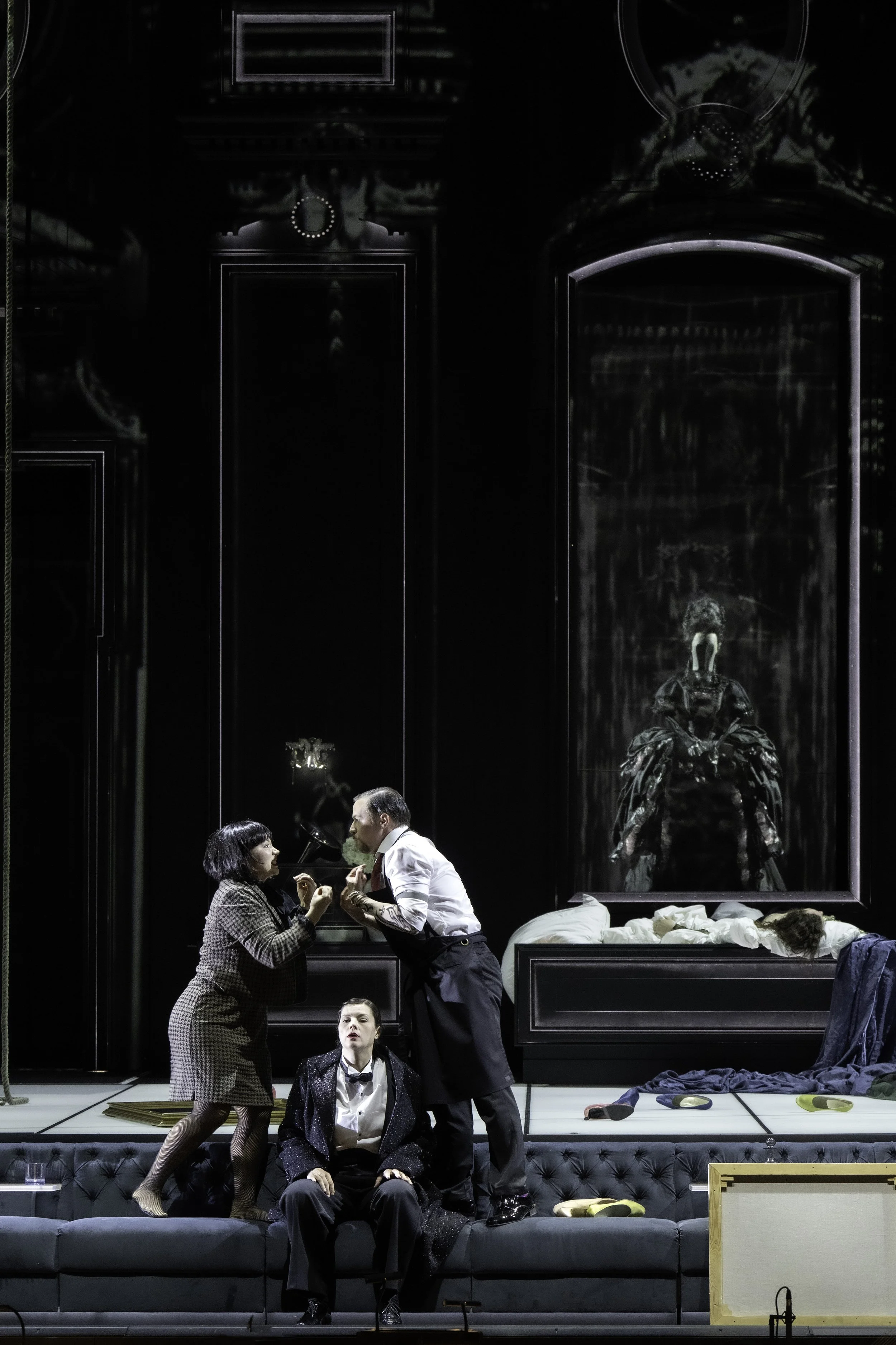 A theatrical performance on stage depicting a surreal domestic scene with people dressed in vintage clothing. Two individuals are standing and engaging intensely, while another person is sitting on a sofa with a dramatic expression. The stage backdro