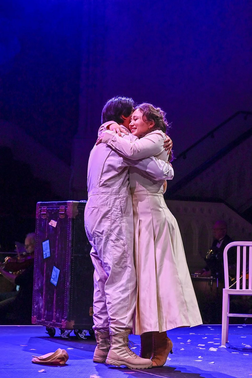 Two people hugging warmly on stage during a theatrical performance, with a dark background and musical instruments in the background.