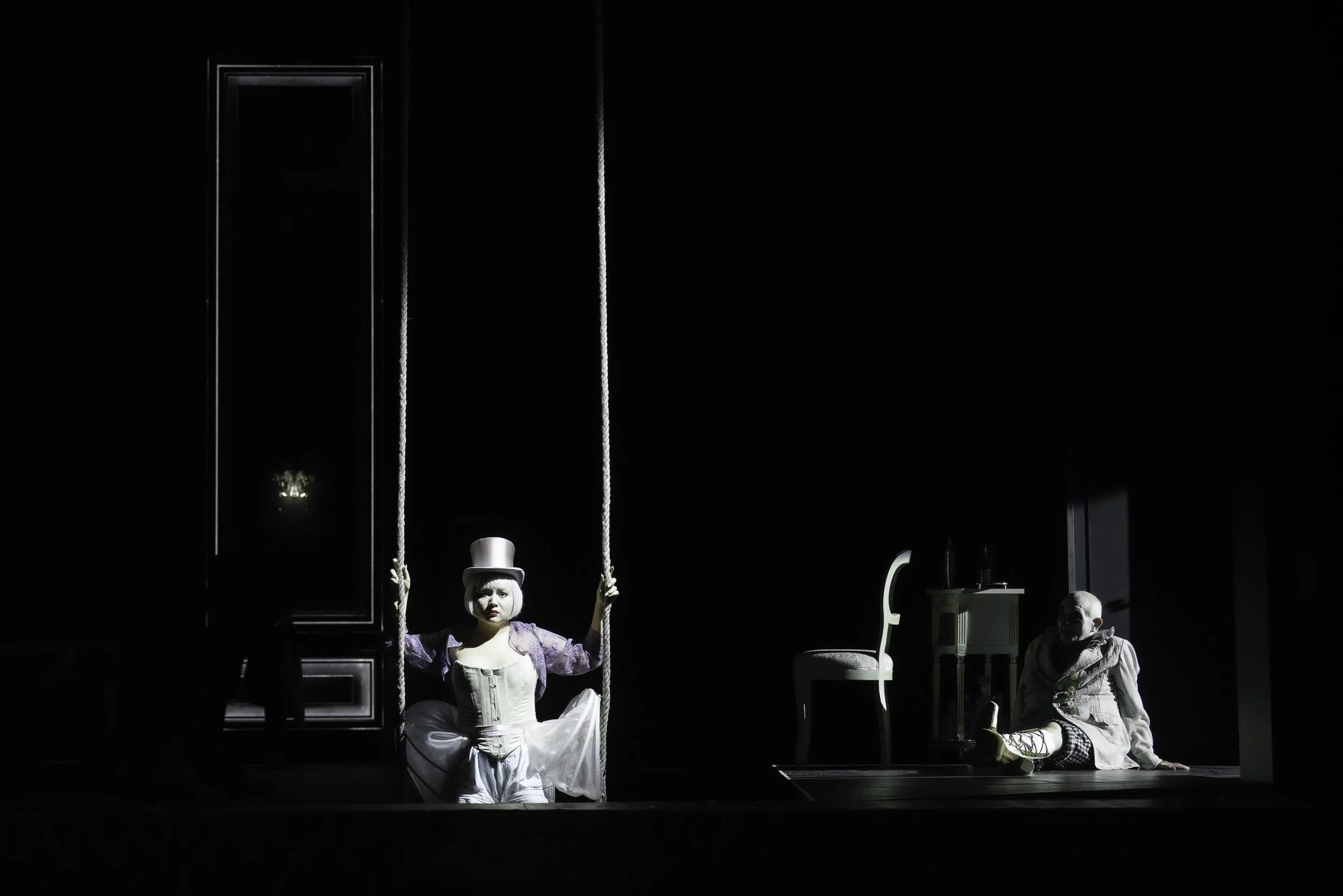 A theatrical stage scene with four actors, including a woman in a costume sitting on a swing, and a man sitting on the floor, with dark background and minimal lighting.