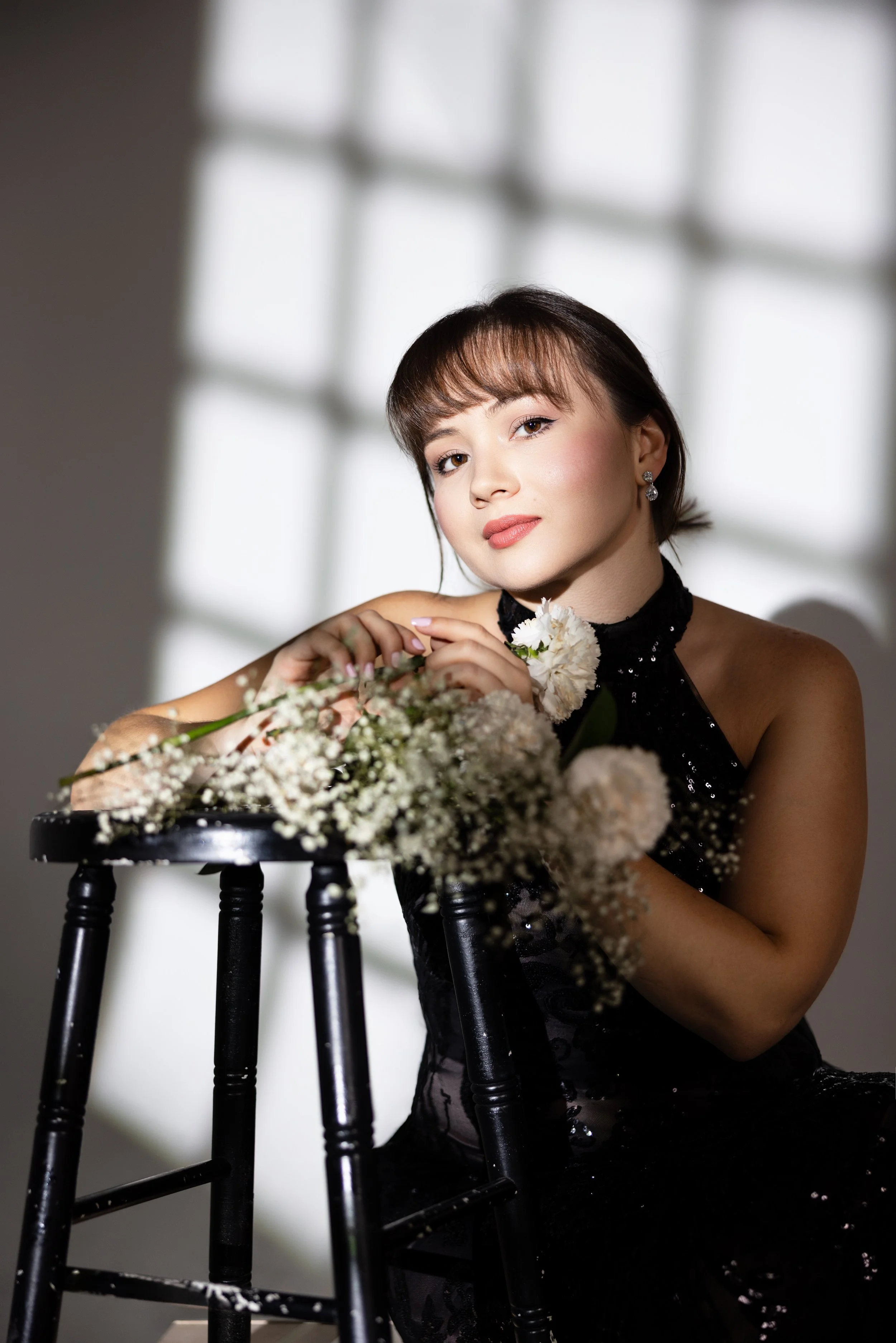A woman with brown hair, wearing earrings and a black sequined dress, sitting behind a small black table with white flowers, in front of a window with white blinds.