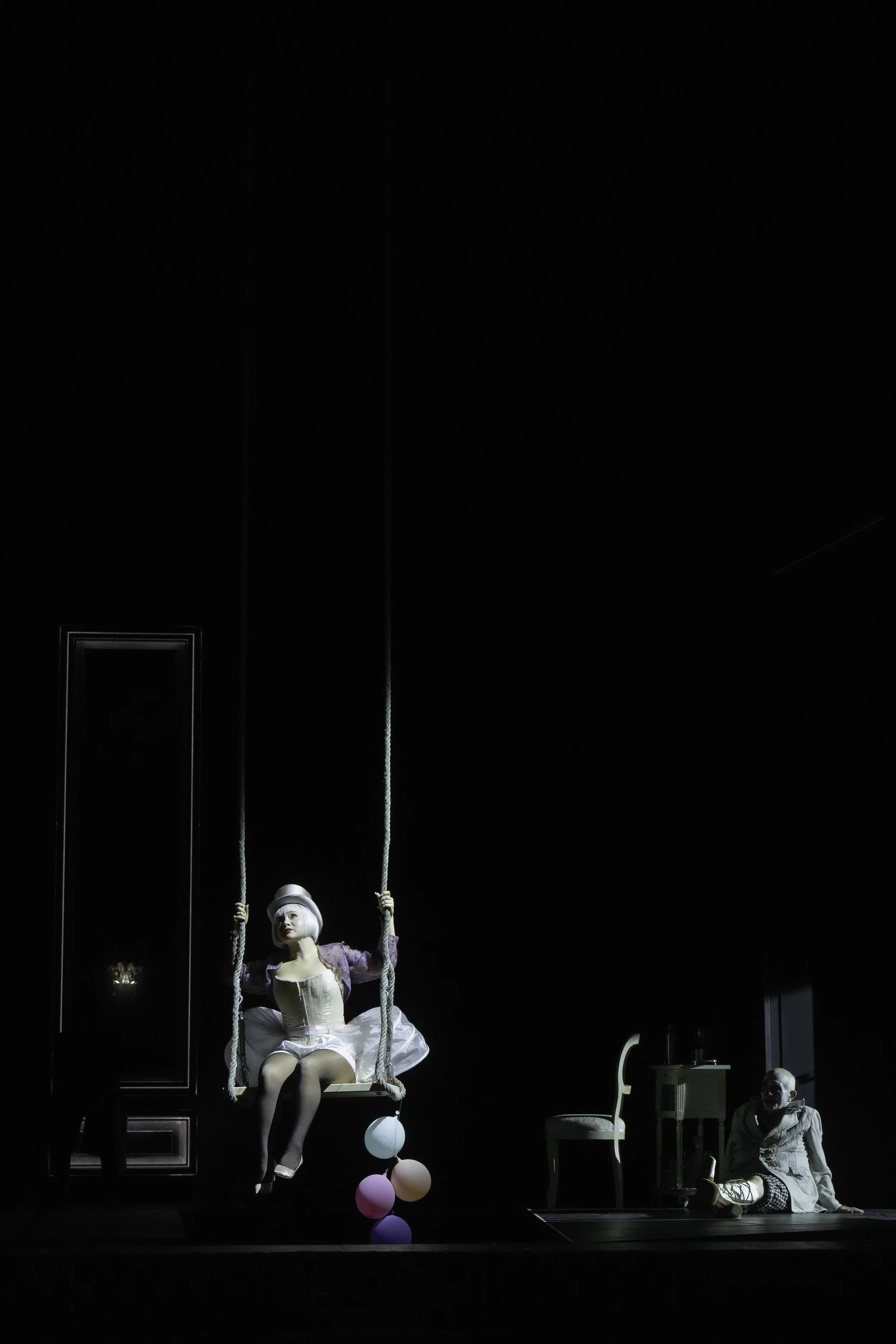 A theatrical performance with a woman dressed as a clown on a swing, wearing a purple top hat and a ruffled white dress, with balloons hanging from the swing. A man in costume sits on the ground to the right, watching the scene. The setting is a dark