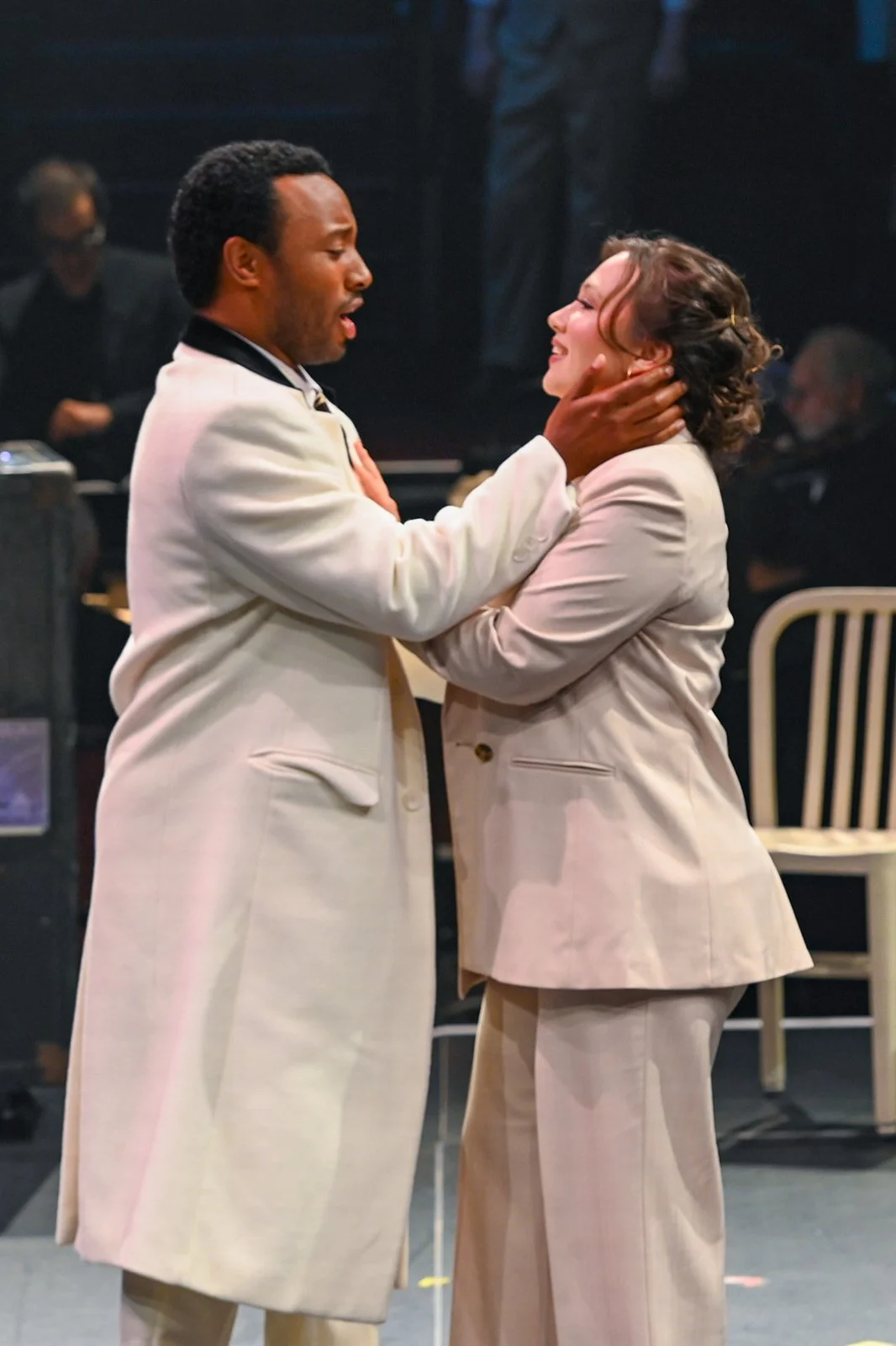 Two people dressed in beige suits are hugging and smiling, with a dark background and a person playing a piano in the distance.