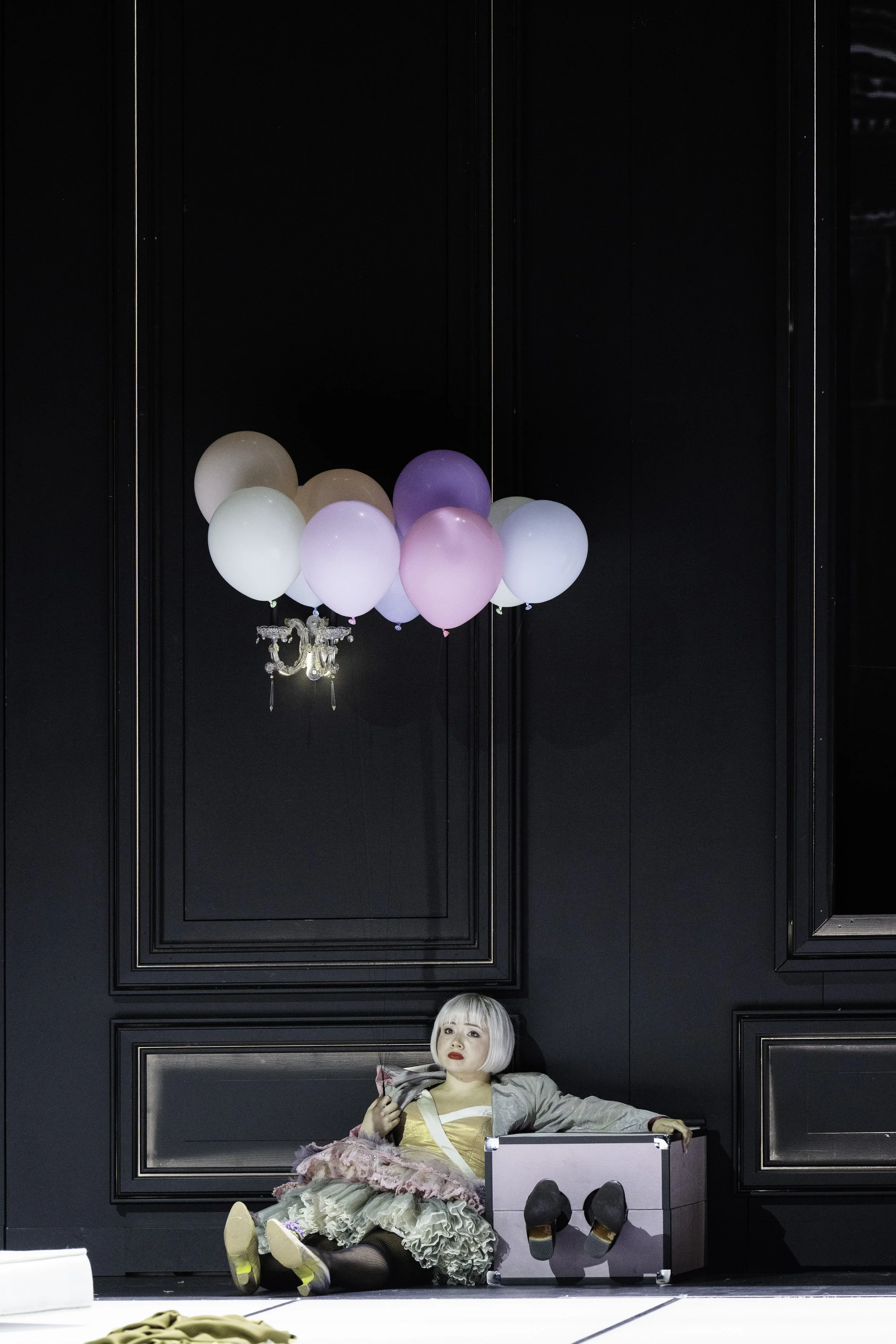 A person with short blonde hair and red lipstick sitting on the floor leaning against a black wall, wearing a layered skirt, black fishnet stockings, and a shiny silver jacket, with various balls and balloons hanging above.