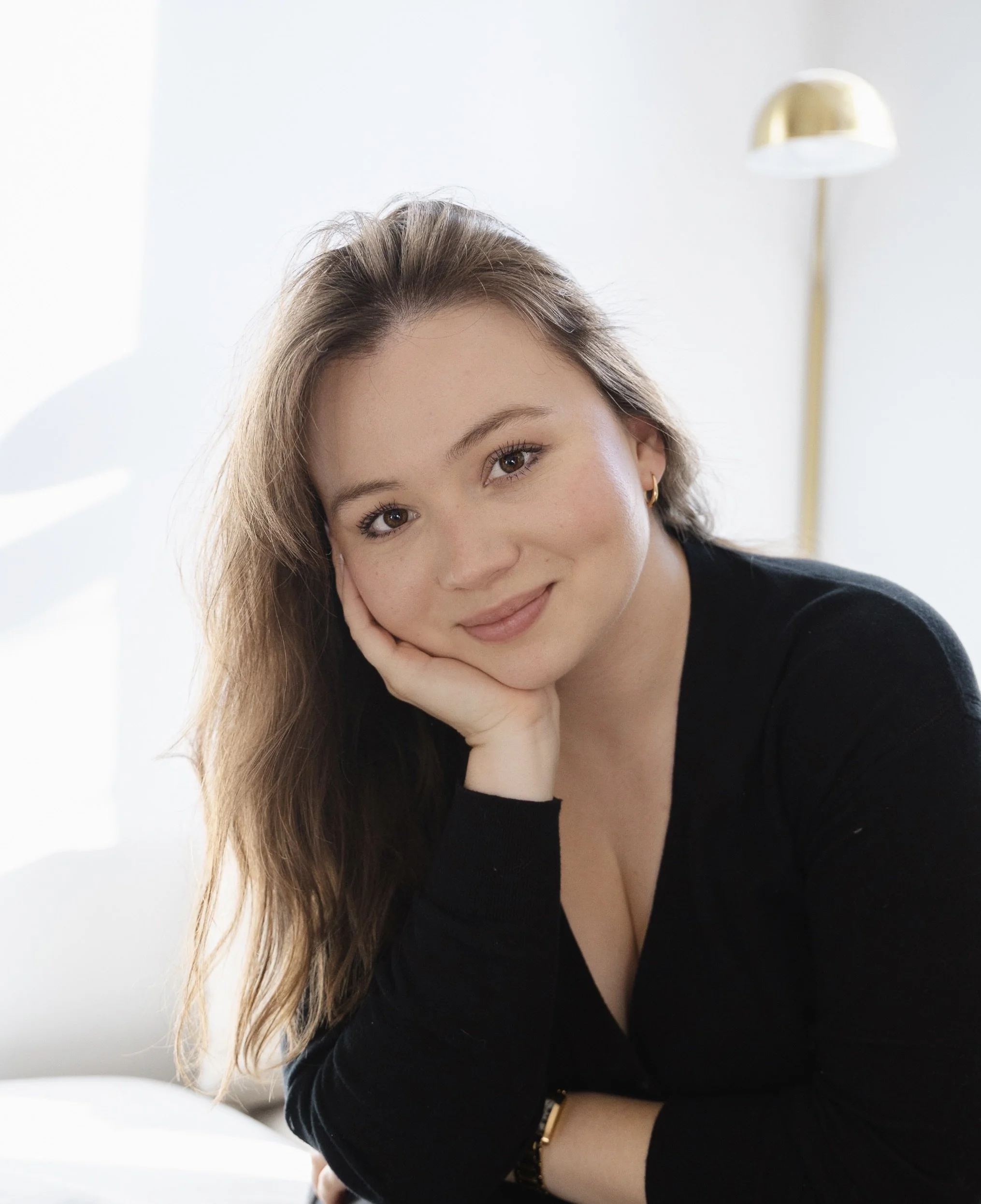 A woman with light skin and long brown hair resting her head on her hand, smiling gently, in a bright, minimalistic room with a white wall and a modern floor lamp in the background.