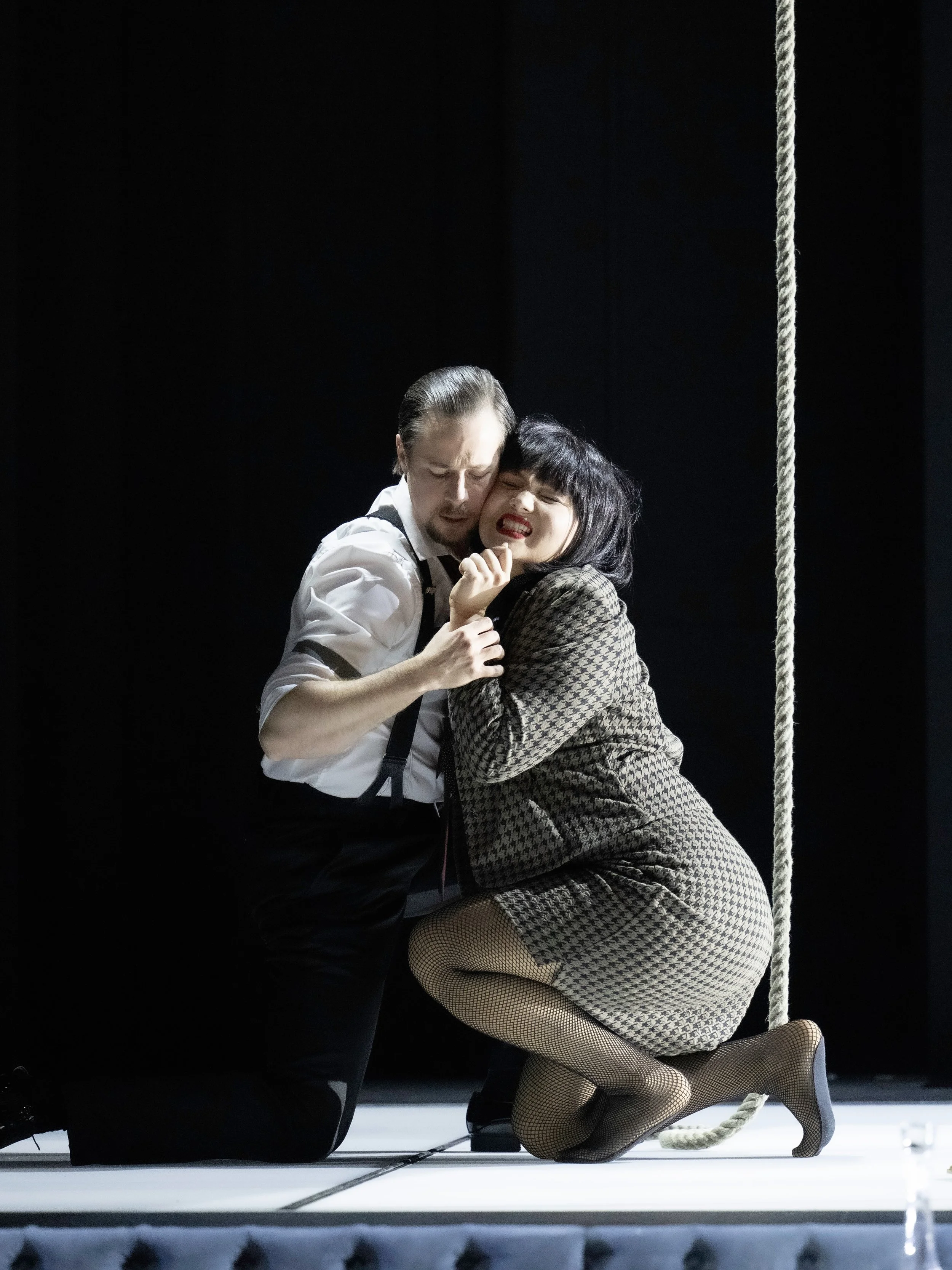 A man and woman in vintage-inspired clothing are engaged in a passionate embrace on a swing. The woman is wearing a houndstooth patterned dress, fishnet stockings, and high heels. The man is in a white shirt with suspenders. The scene is set against 