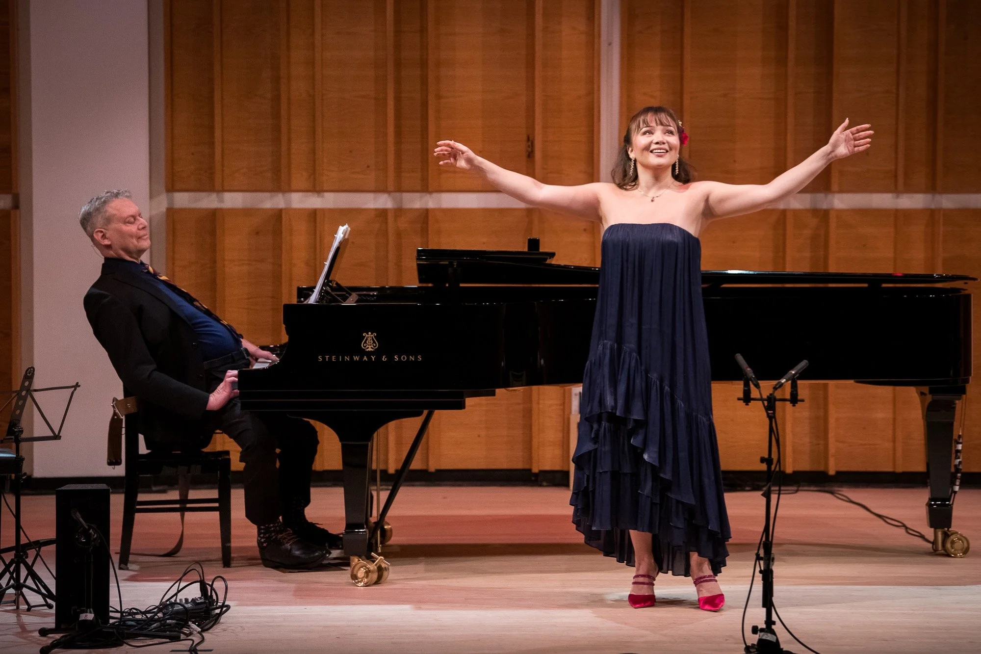 A woman in a navy blue dress and red heels is performing on stage with her arms outstretched. A man in a black suit is playing a grand piano beside her. The stage has wood-paneled walls.