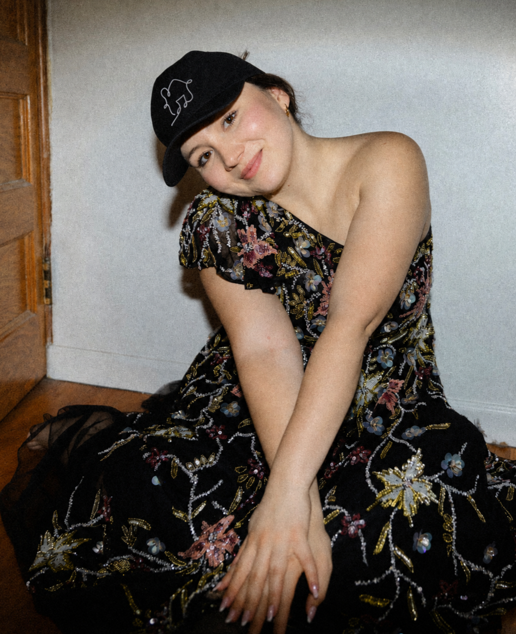 A woman sitting on the floor wearing a floral dress and a black baseball cap with a white embroidered outline of a dog. She is smiling softly, with her head tilted and arms crossed over her knees, outside a door with a wooden frame.