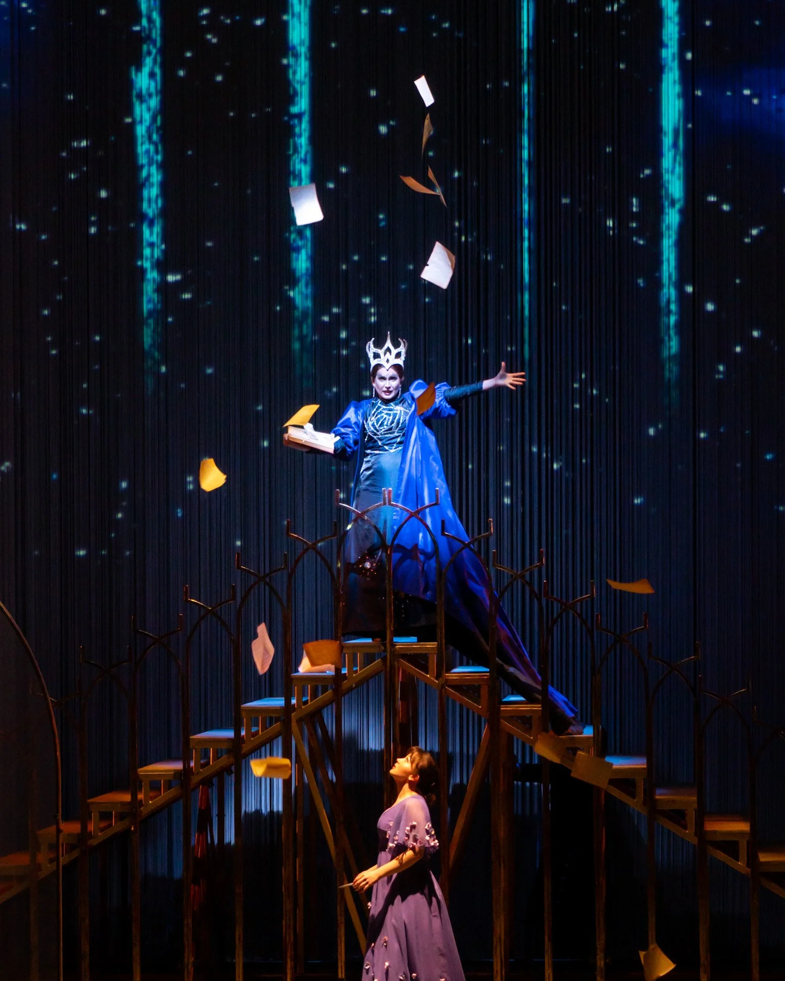 A theatrical stage scene with two actresses in period costumes. One actress, standing on a wooden staircase, reaches out with her arms open, while the other actress, standing on the stage below, looks up. The stage backdrop features a digital blue st