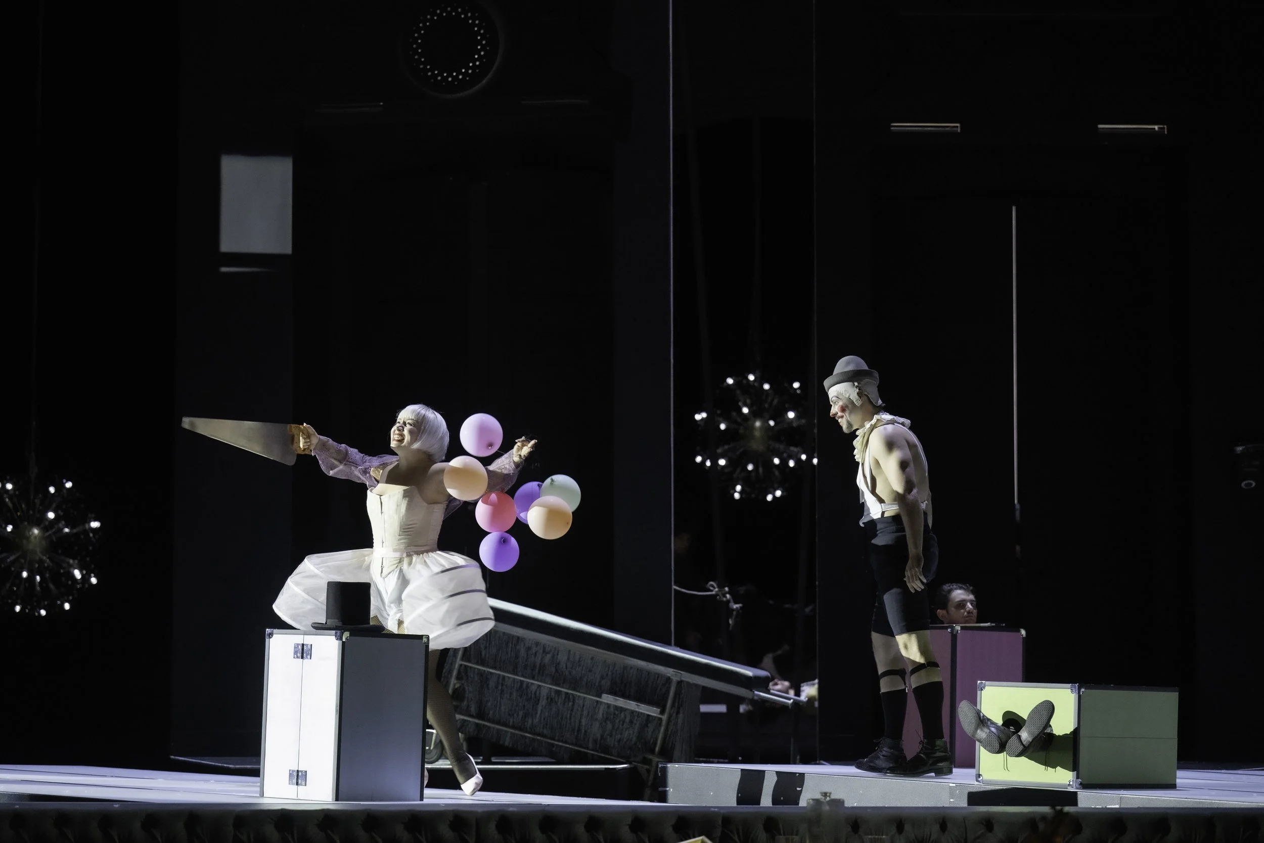 Two performers on stage, one woman in a white dress holding balloons and a man in a clown costume and gray hat, with Christmas trees decorated in the background.