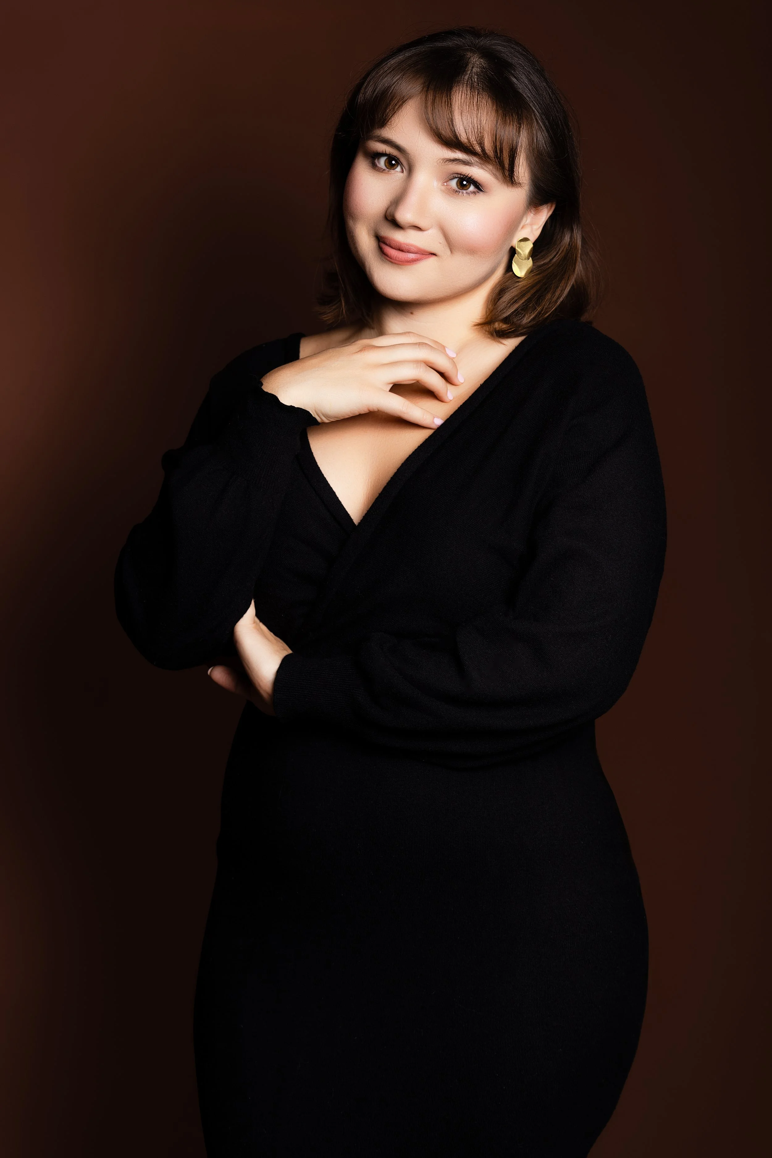 A woman with short brown hair wearing a black dress and gold earrings, posing with a soft smile and hand near her collarbone against a dark brown background.