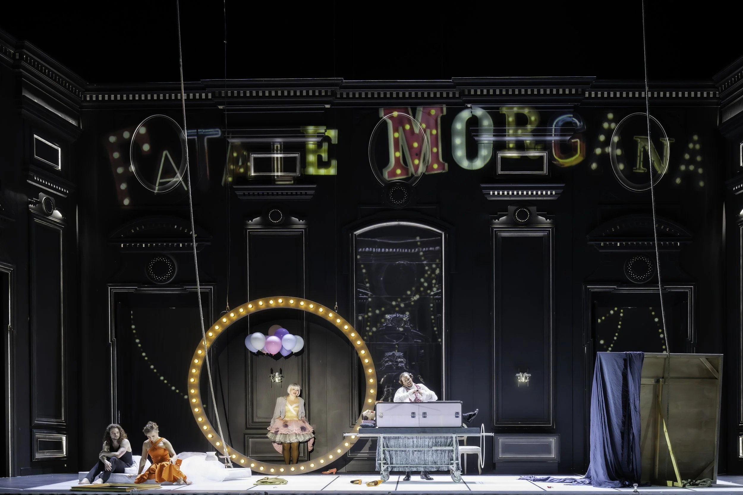 A theatrical stage with a dark ornate backdrop, illuminated circular frame, and people dressed in costumes performing. Balloons are hanging and scattered on stage, indicating a lively performance or rehearsal.