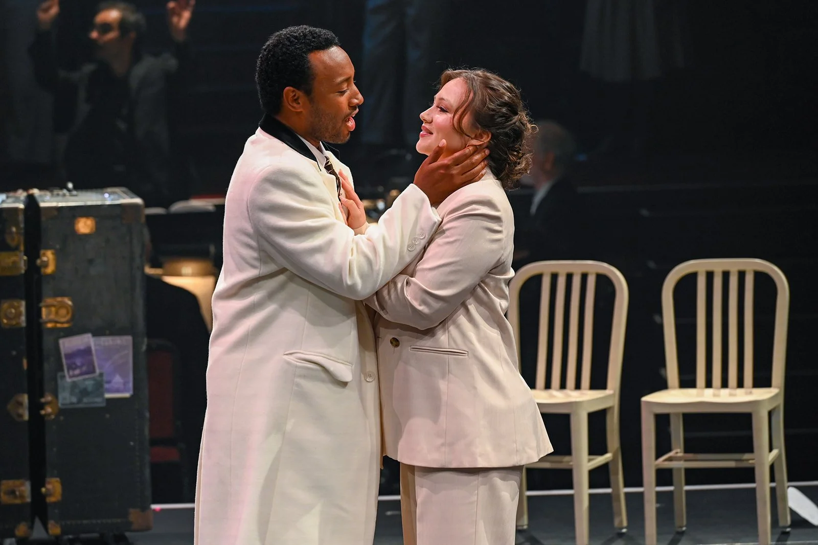 Two actors in white suits are on stage, embracing each other affectionately during a theatrical performance.