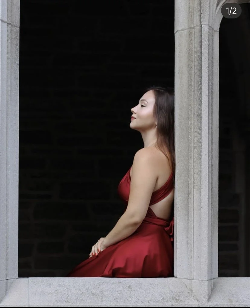 A woman in a red dress sitting by a stone window frame with a dark background.
