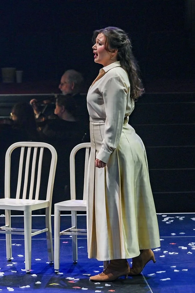 A woman singing on stage with two white chairs in front of her, wearing a beige outfit and brown high heels, with confetti on the floor and an orchestra in the background.