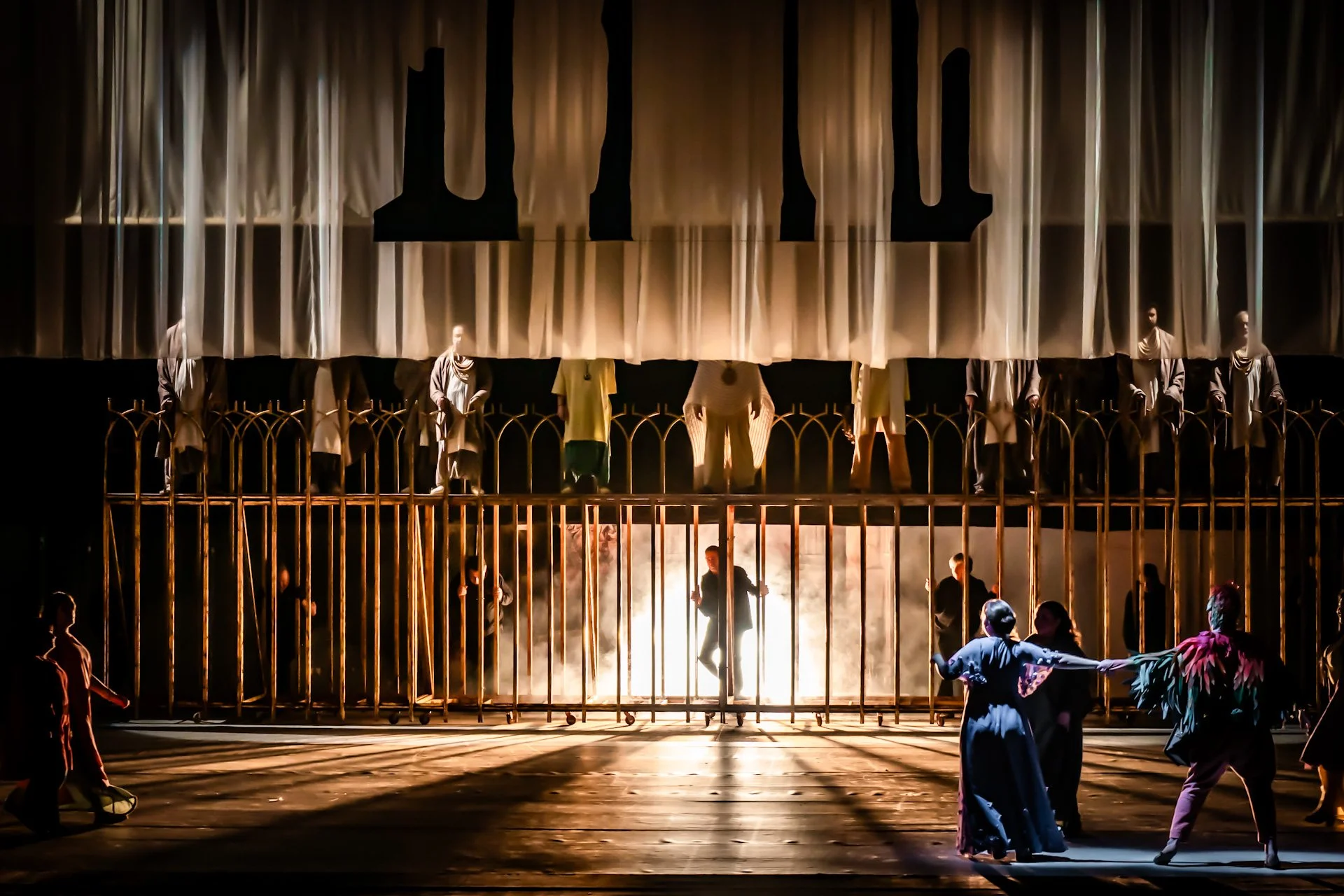 The image shows a theatrical stage scene with people acting and standing behind a large gold-colored fence. There is a large, partially lifted curtain with a dark backdrop and some lighting effects, creating a dramatic atmosphere. Several actors are 
