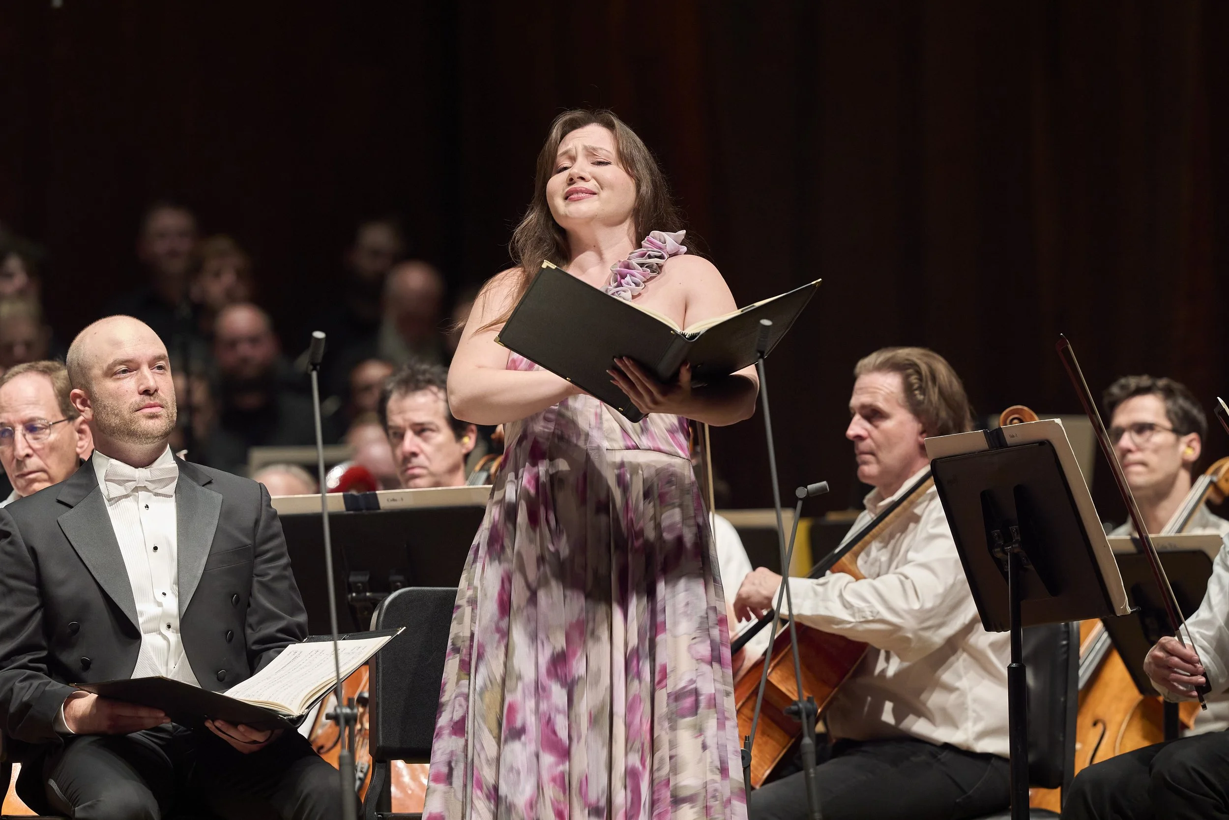 A woman in a floral dress singing with an orchestra during a performance.