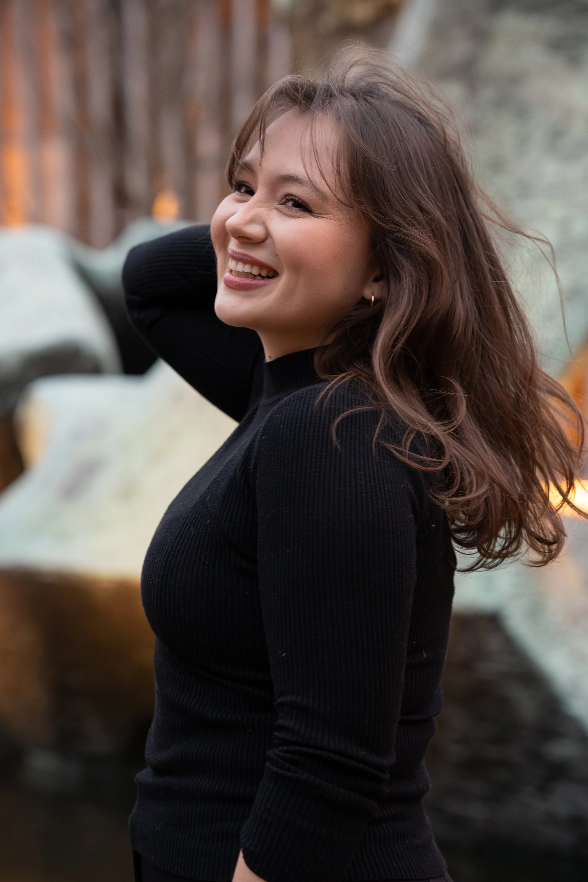 A woman with long wavy brown hair smiling while posing outdoors, wearing a black long-sleeve top and earrings, with a blurred natural background.