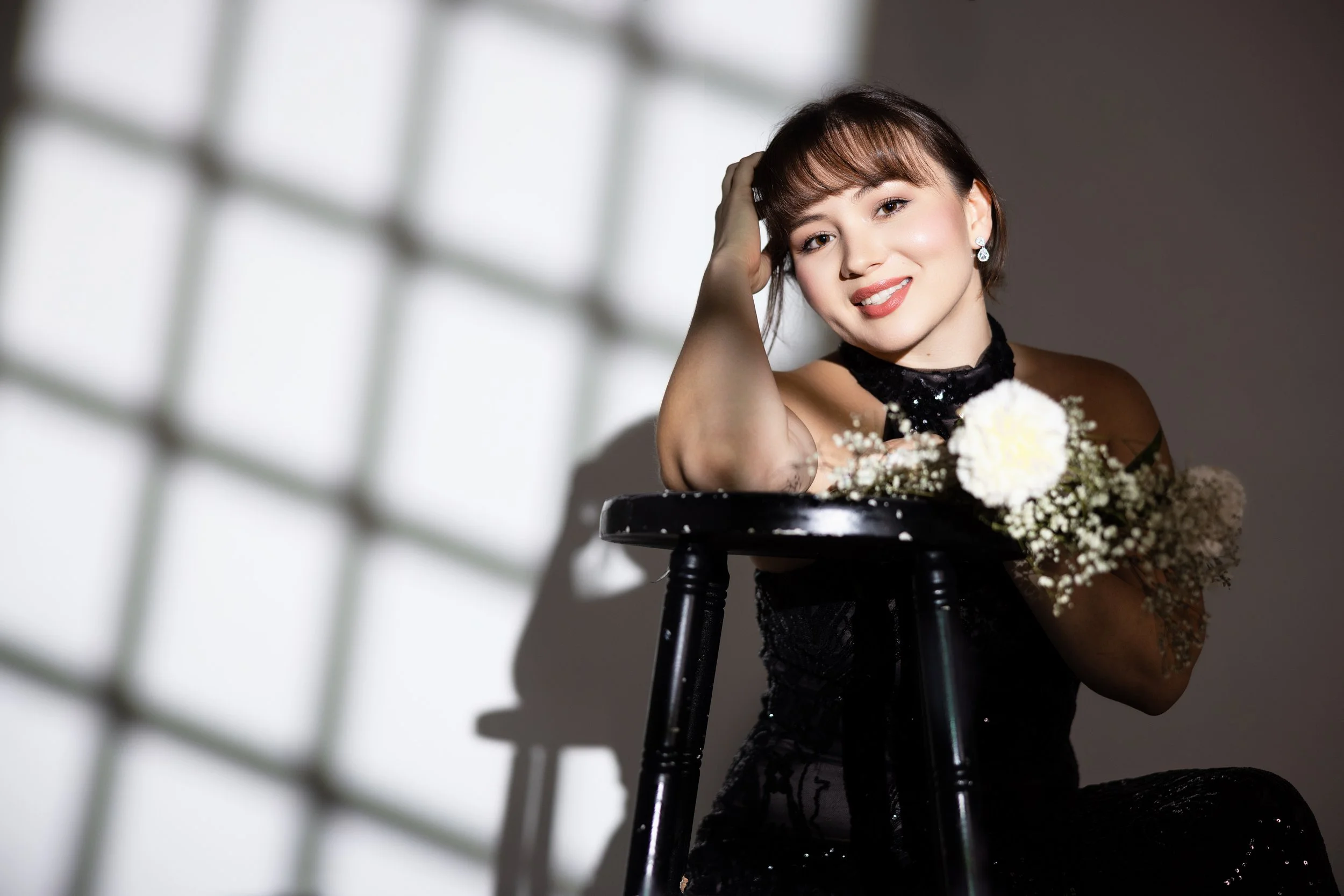 A woman with short brown hair, wearing earrings and a black dress, sits at a black table with a bouquet of white flowers, smiling at the camera in a studio setting with soft lighting.