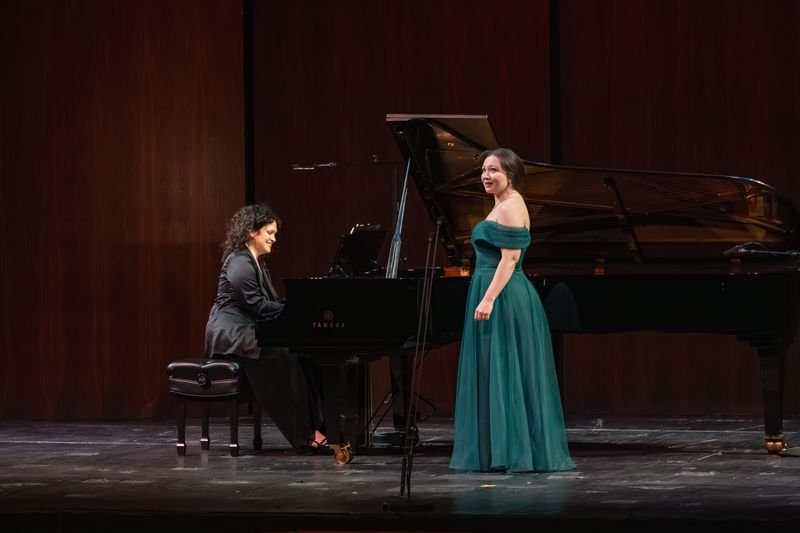 A woman in a green gown singing on stage with a pianist playing a grand piano beside her.