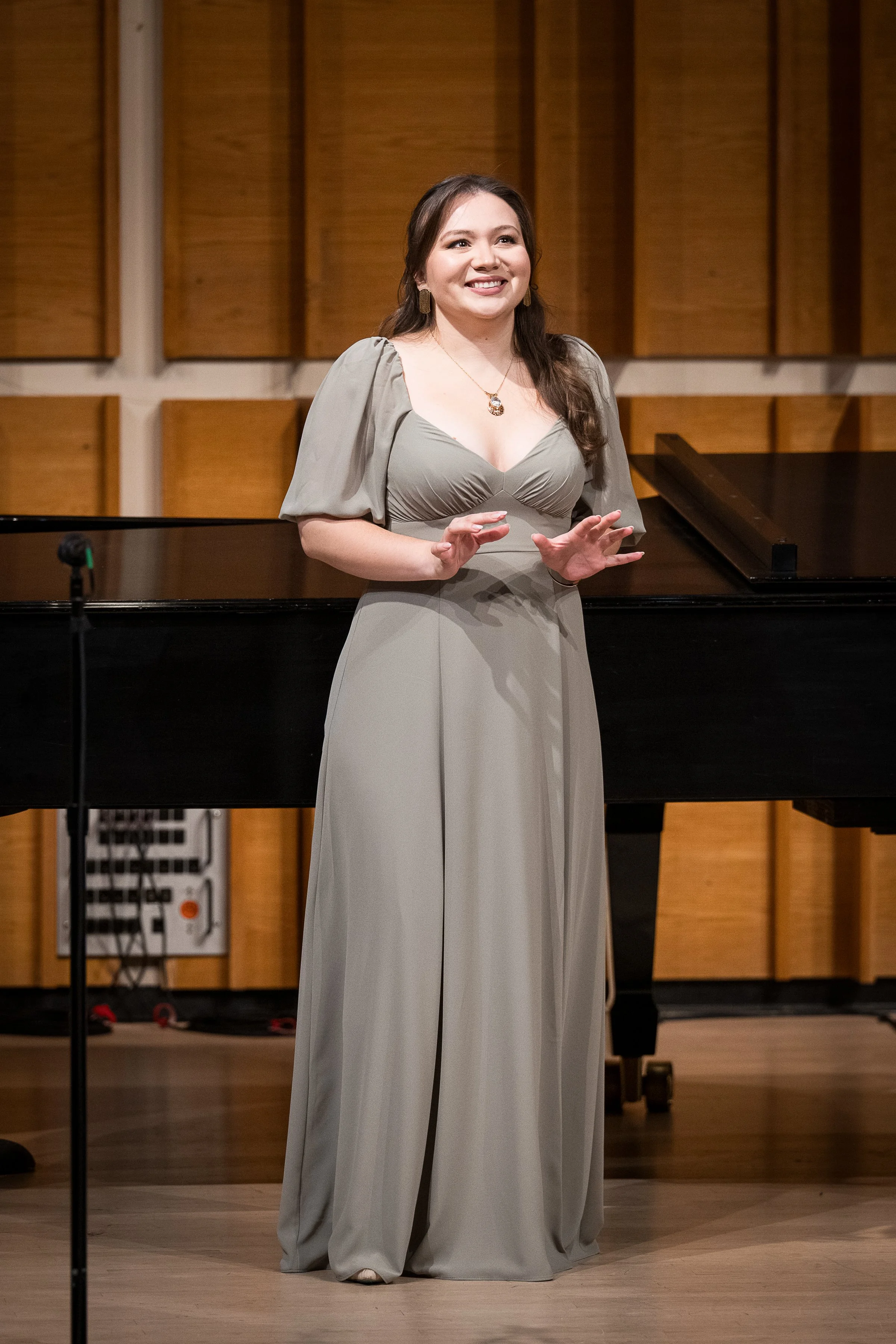 A woman in a long, elegant taupe dress stands on stage in front of a wooden panel background, smiling and gesturing with her hands.