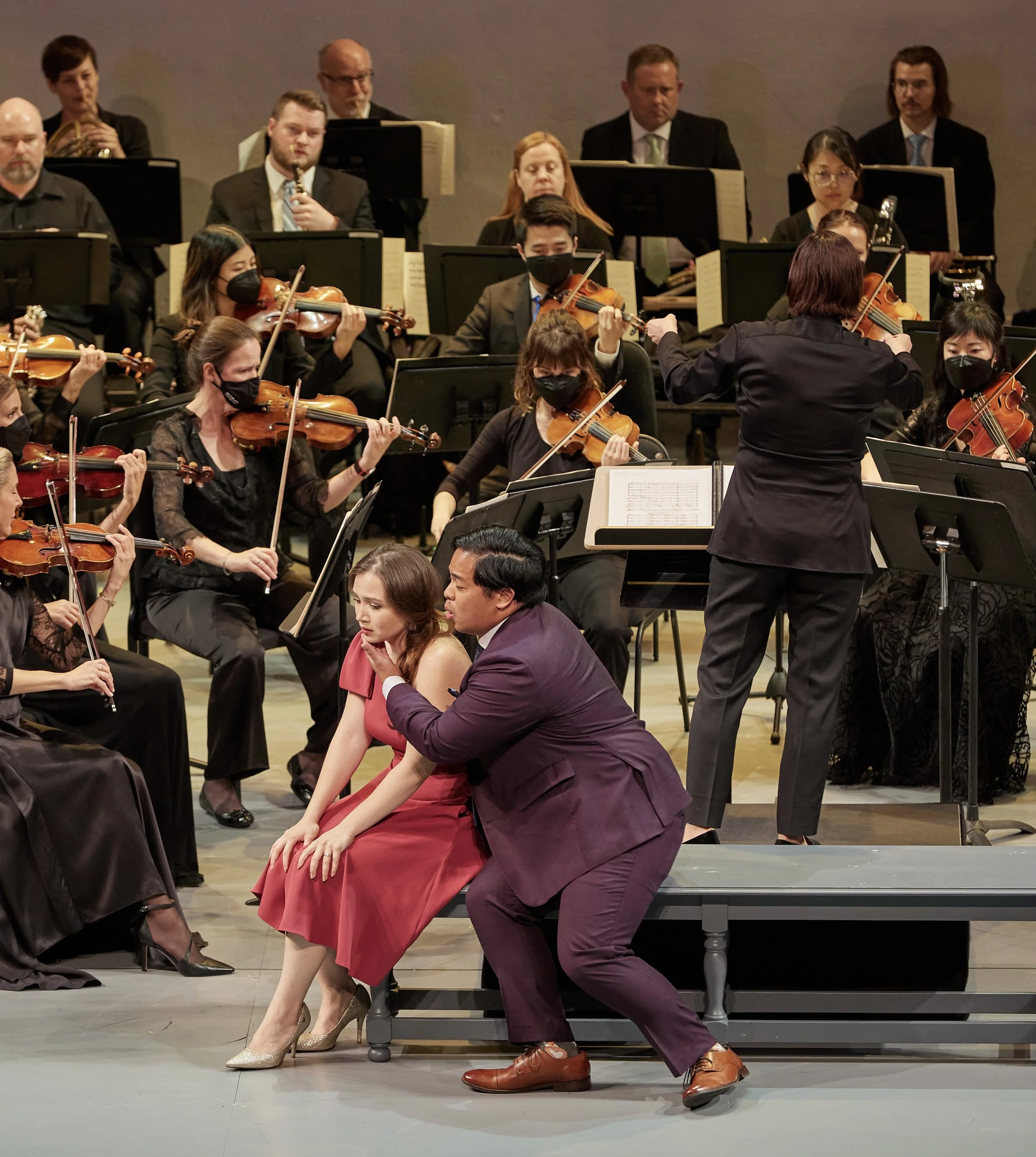 Orchestra performing on stage with conductor in foreground, musicians playing violins, violas, similar instruments, some wearing face masks, in a concert hall setting.
