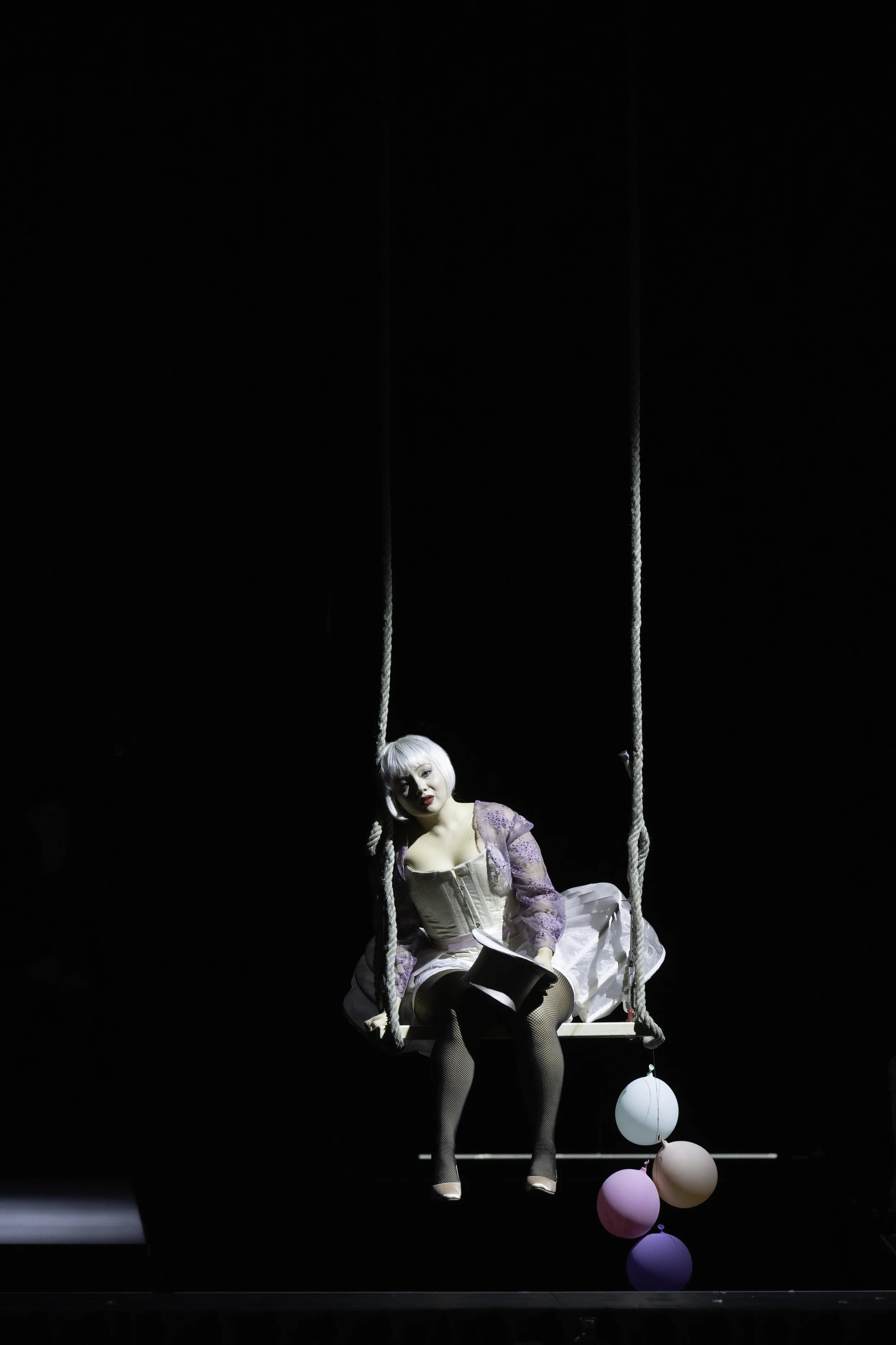 A woman with a short platinum blonde wig and makeup, dressed in vintage-inspired clothing, is sitting on a swing on a dark stage. She is holding a black top hat and surrounded by pastel-colored balloons hanging below the swing.