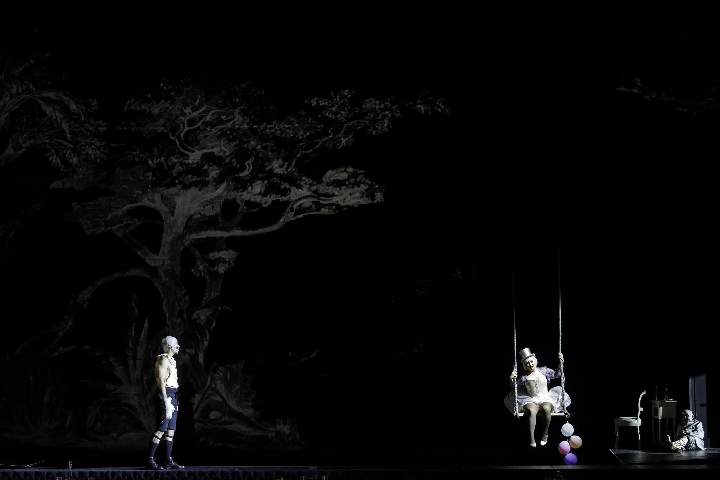 Stage scene with performers, including a woman on a swing and a man standing nearby, set against a dark background with a large tree depicted in the backdrop.