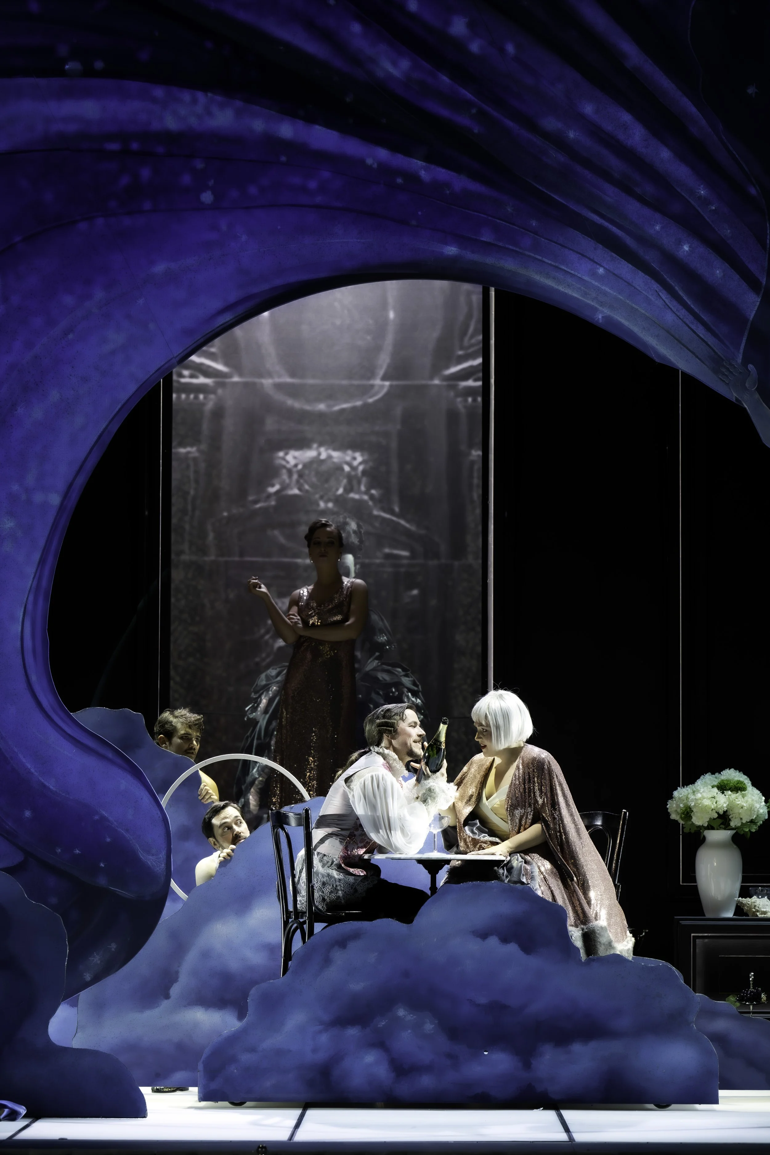 Actors performing on stage with a blue set design, including clouds and moon motifs. Central female actor in a sparkly dress stands behind a seated man in a white shirt, with other actors around them.