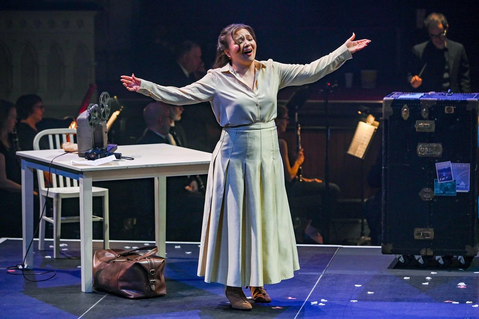 Woman singing on stage with arms outstretched, surrounded by musicians, with a suitcase and desk nearby.