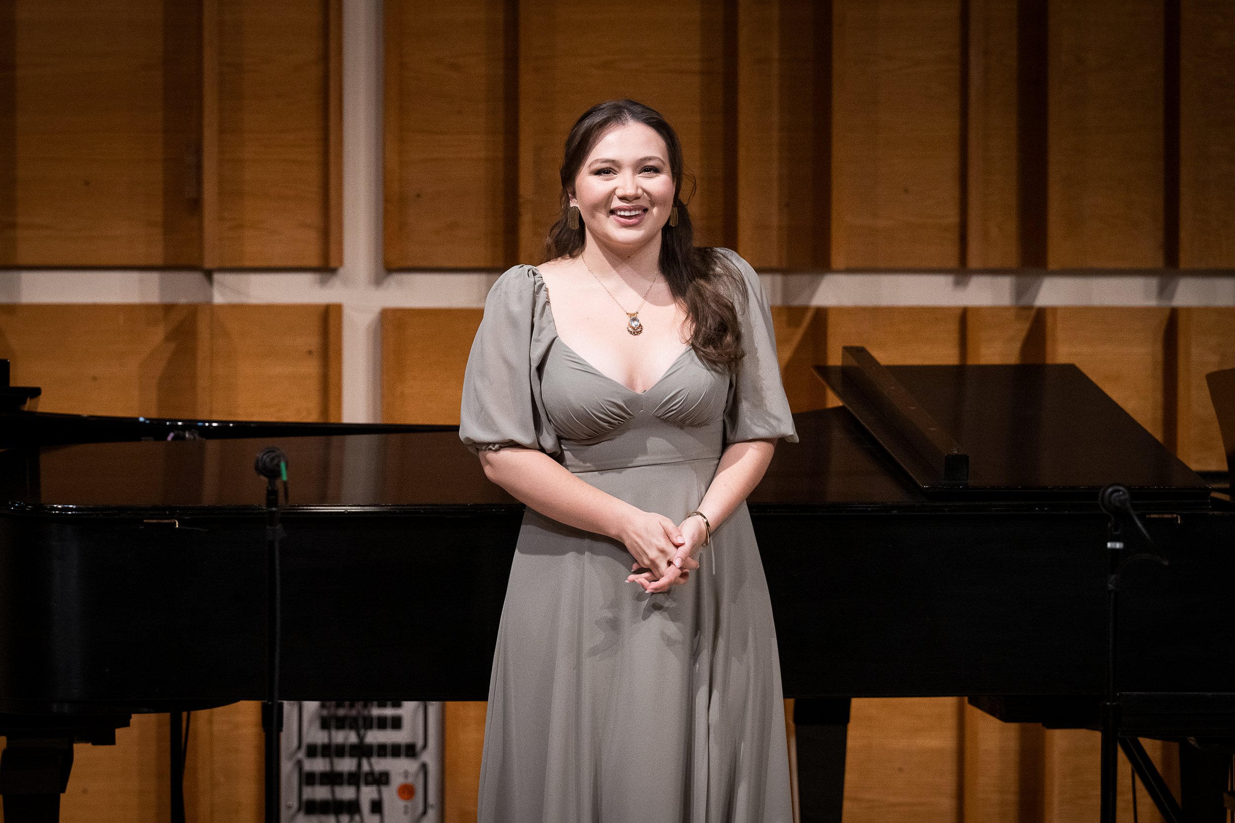 A woman in a gray dress standing in front of a piano in a room with wooden acoustic panels on the wall.
