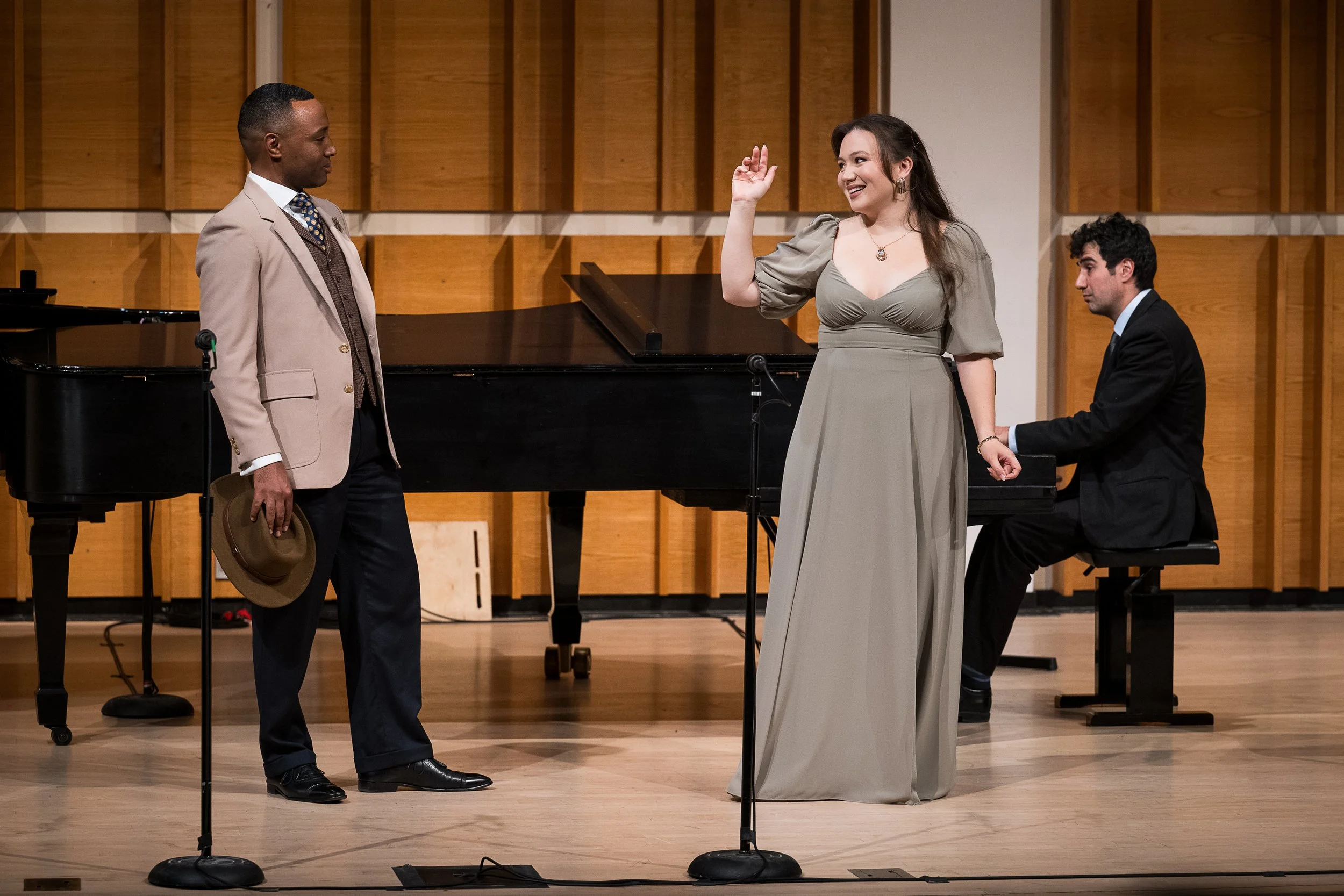 Three performers on stage in a concert hall. One man in a beige suit holds a hat, a woman in a gray dress is smiling and gesturing with her hand, and a man in a black suit is seated at a piano.