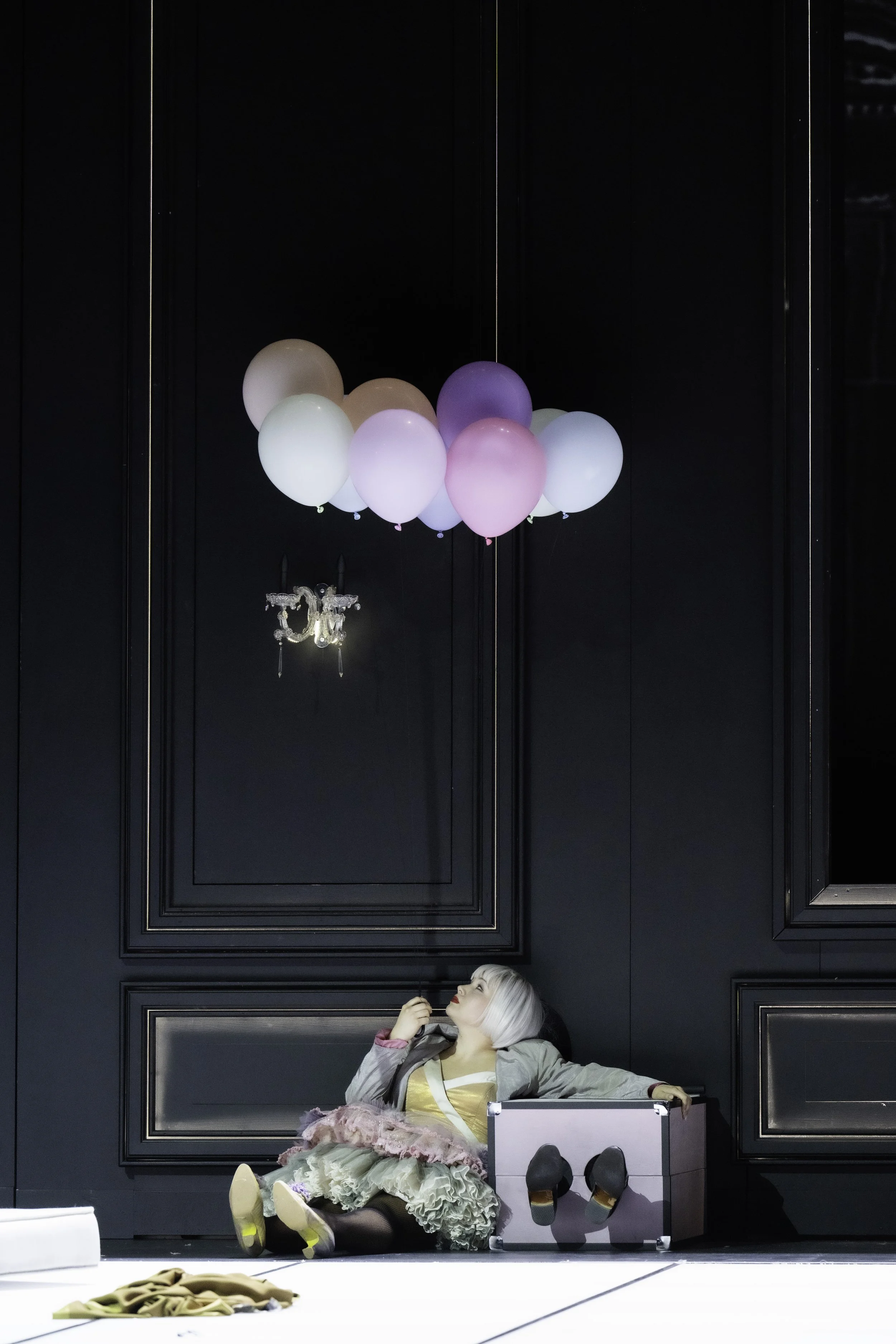 A person with short platinum blonde hair and colorful makeup lying on the floor beneath a dark paneled wall. They are dressed in a layered, ruffled skirt, a gray jacket, and tan shoes, and are playing with pink and purple balloons suspended from the 