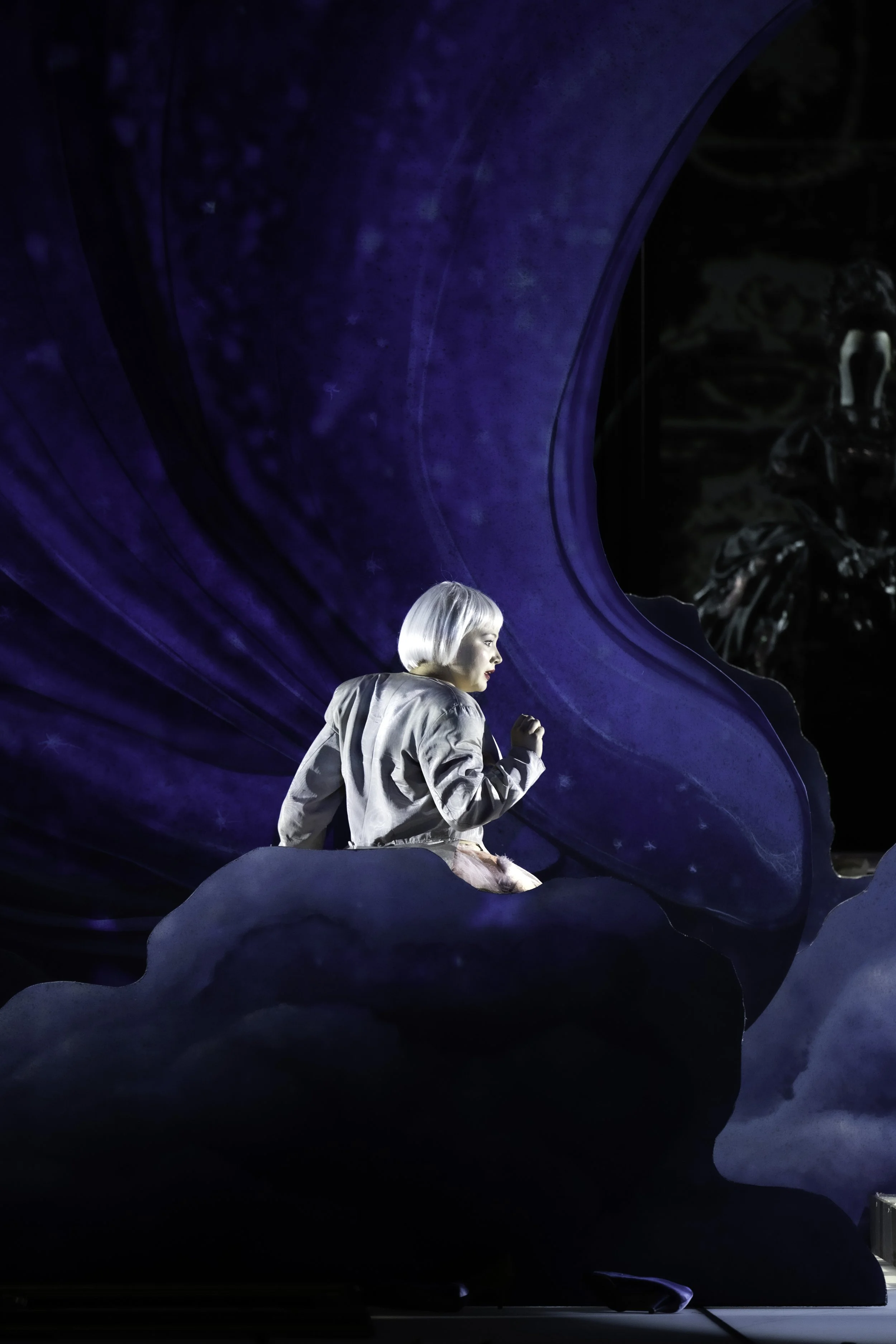 A woman with platinum blonde hair wearing a shiny silver jacket, sitting among large blue floral-like structures, with a dark background and a skeleton visible in the distance.