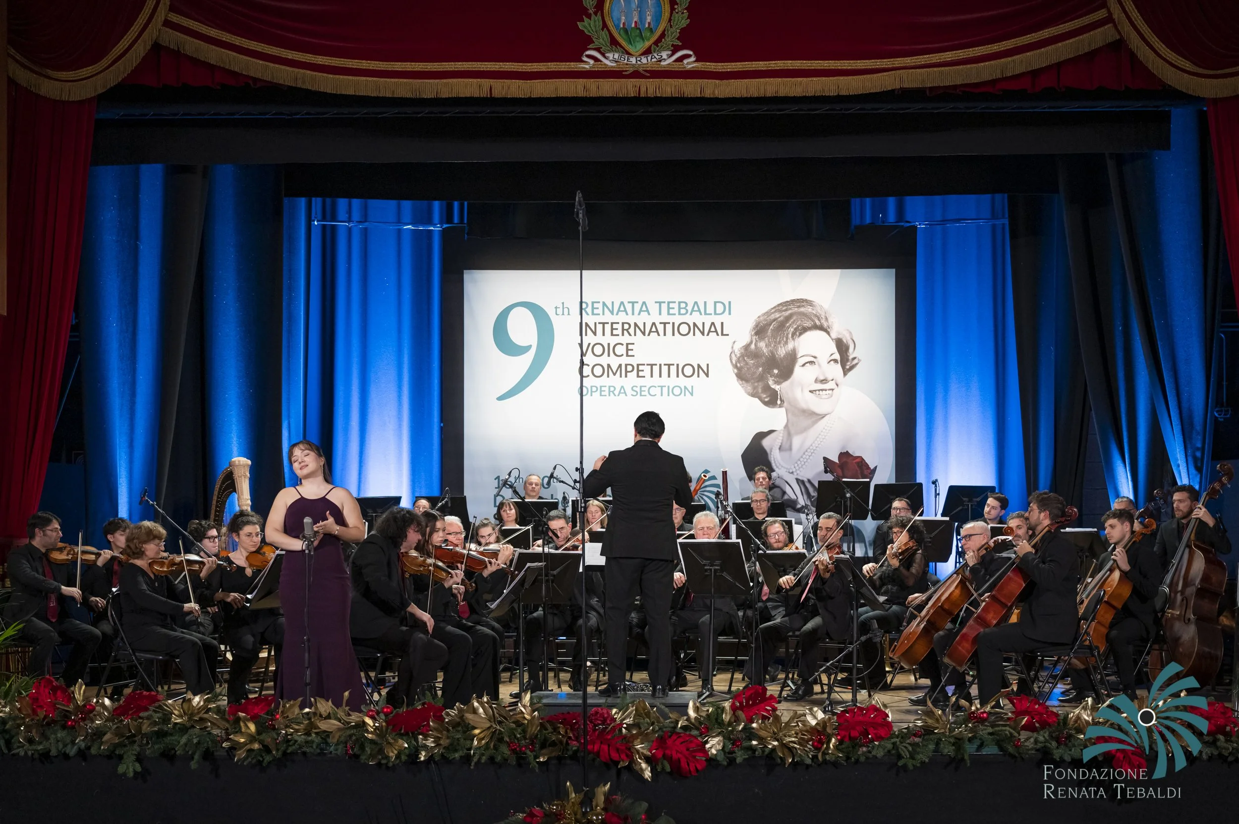 An orchestra performing on a stage decorated with holiday-themed flowers and greenery. A woman in a purple dress stands with her hands clasped in front of her, while a conductor leads the orchestra. A large banner behind the orchestra displays a phot