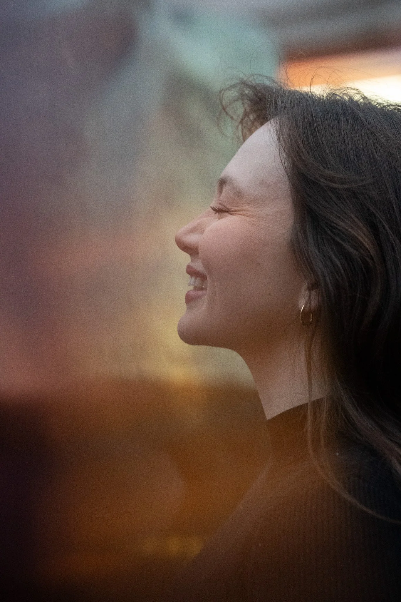 A close-up profile of a woman with dark hair, smiling with her eyes closed, showing her teeth and a small hoop earring.